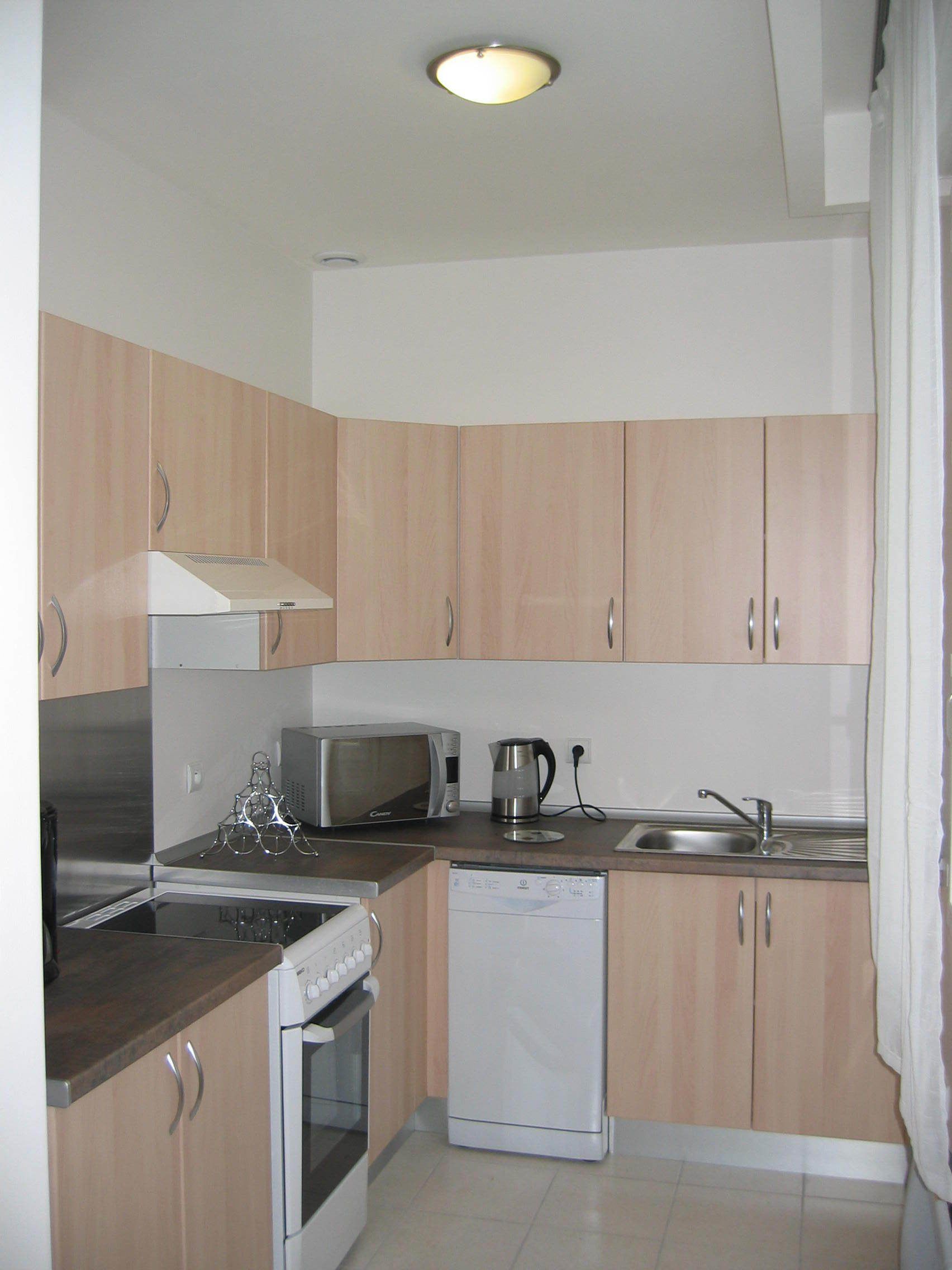 A compact kitchen with wooden cabinets, a microwave, a kettle, a dishwasher, and a stove.