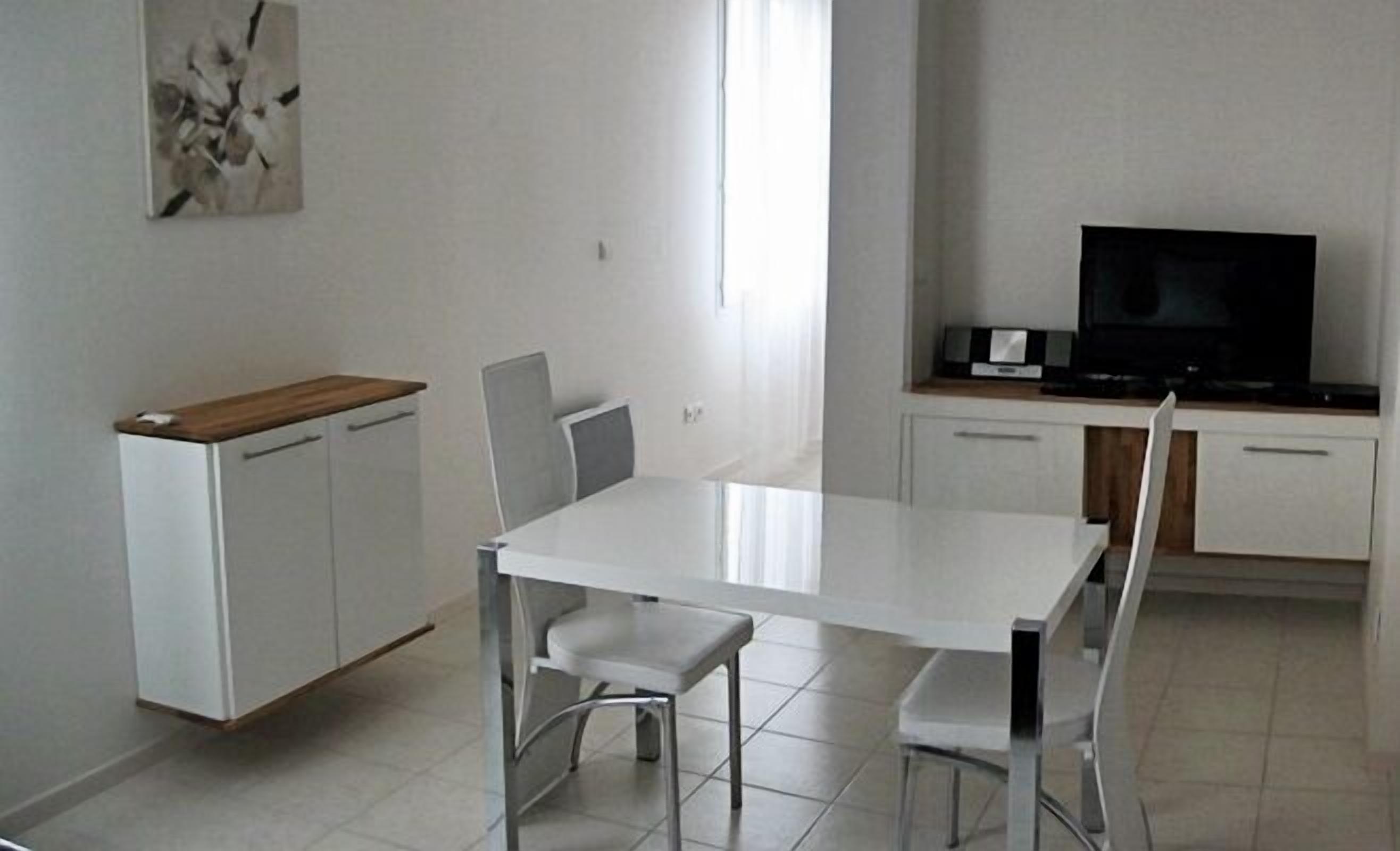 Una habitación moderna y minimalista con una mesa y sillas blancas, una pequeña kitchenette con televisor y un cuadro en la pared.
