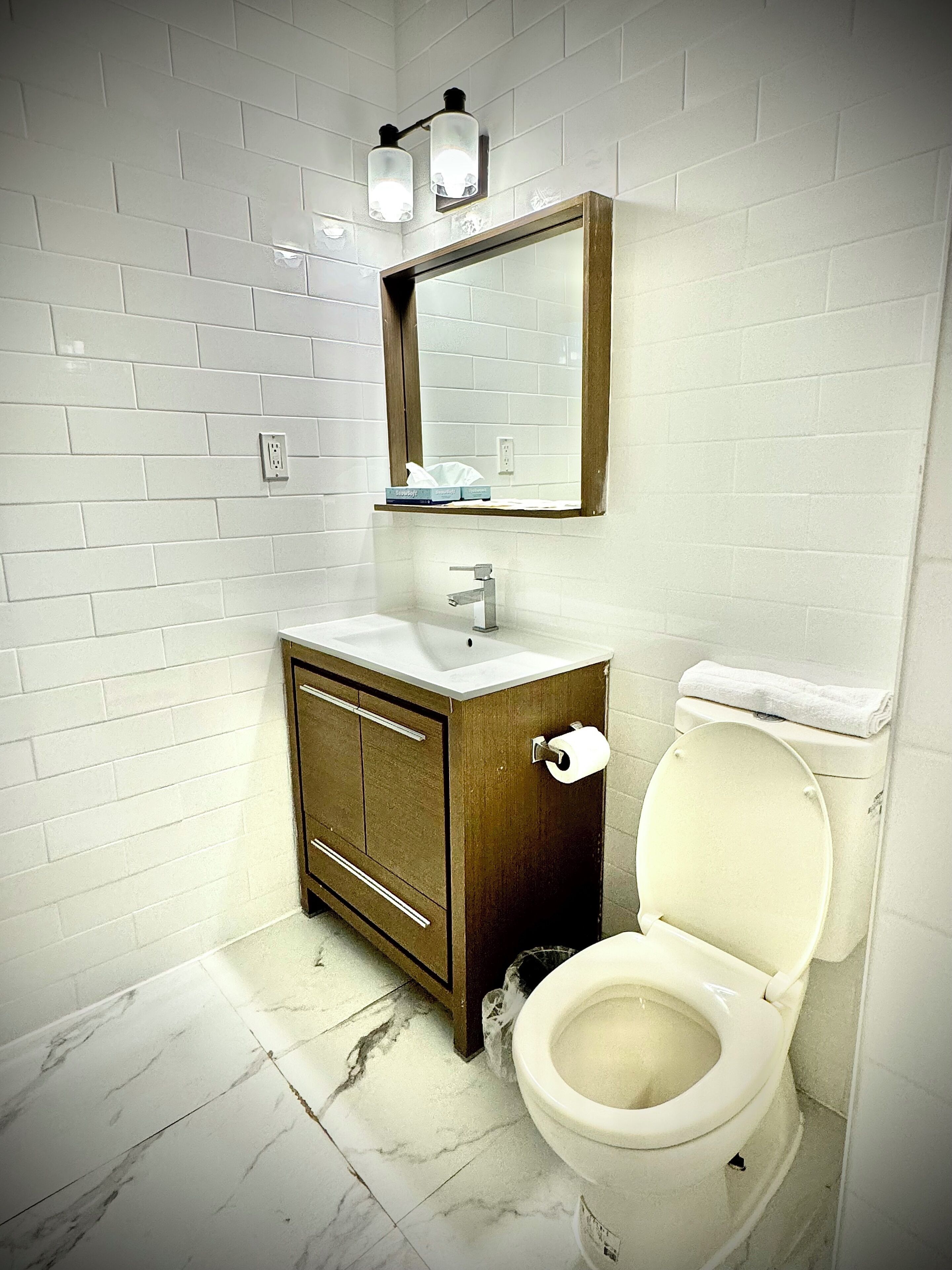 Een badkamer met witte wandtegels, een houten wastafelmeubel, een wit toilet en een spiegel met twee wandlampen.