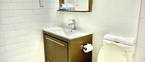 Een badkamer met witte wandtegels, een houten wastafelmeubel, een wit toilet en een spiegel met twee wandlampen.
