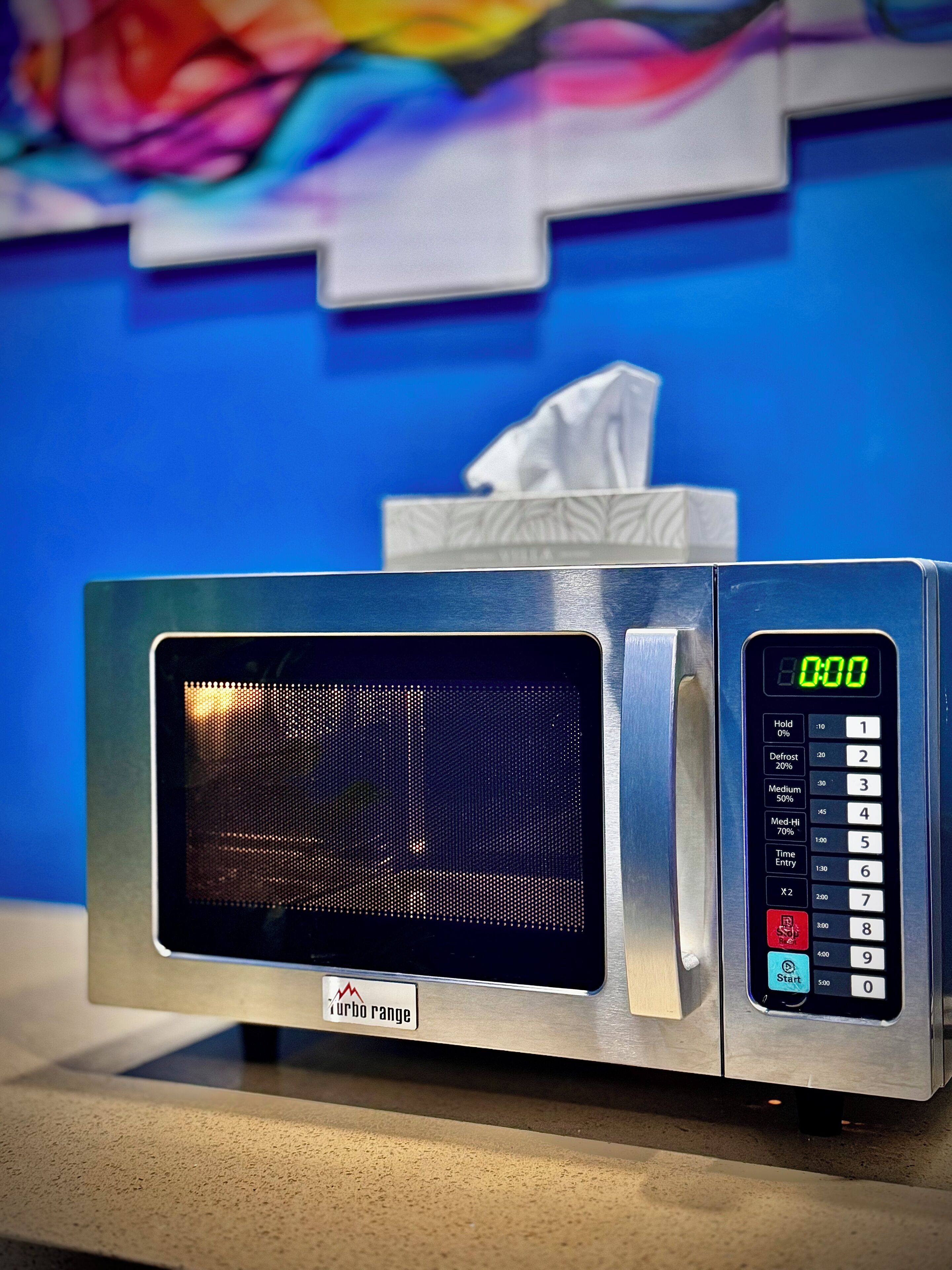 En mikrobølgeovn med digitalt display og kontrolpanel, placeret på en bordplade med blå væg og et maleri i baggrunden.
