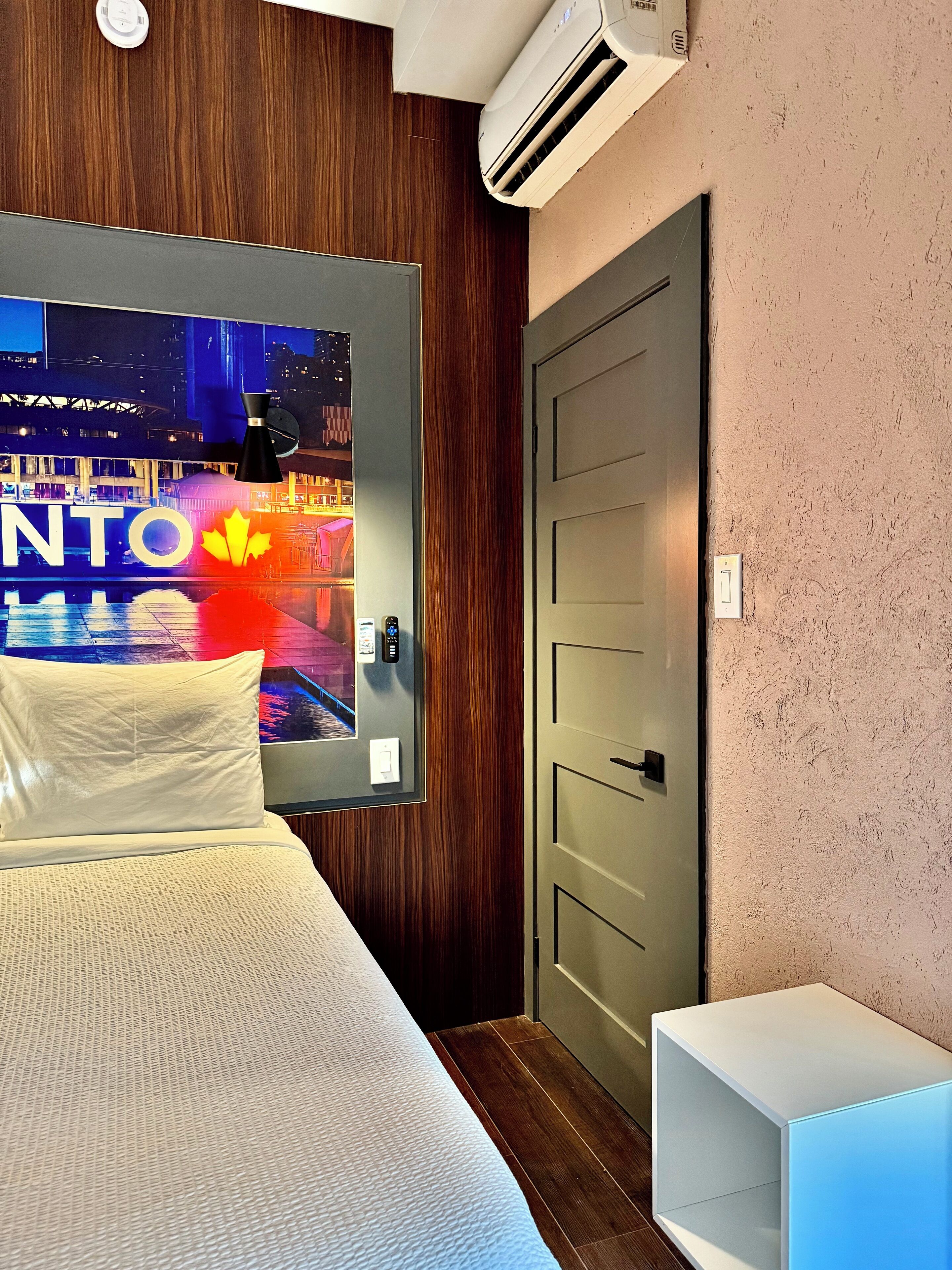 Habitación de hotel con una cama, un cartel con vista nocturna, una mesita de noche, un equipo de aire acondicionado y una puerta.