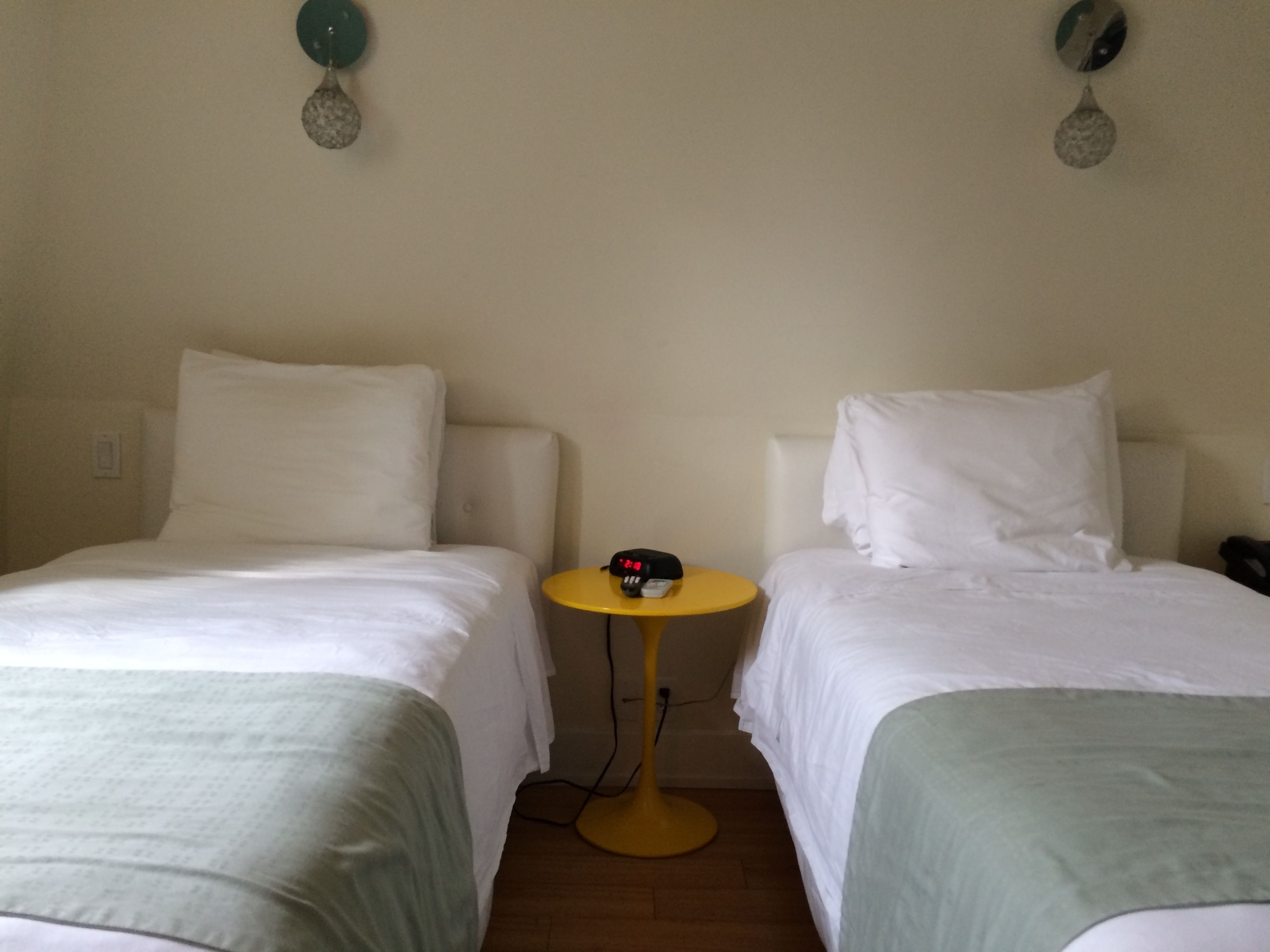 A hotel room with two single beds, a small round table with a clock, and a wall-mounted light fixture.