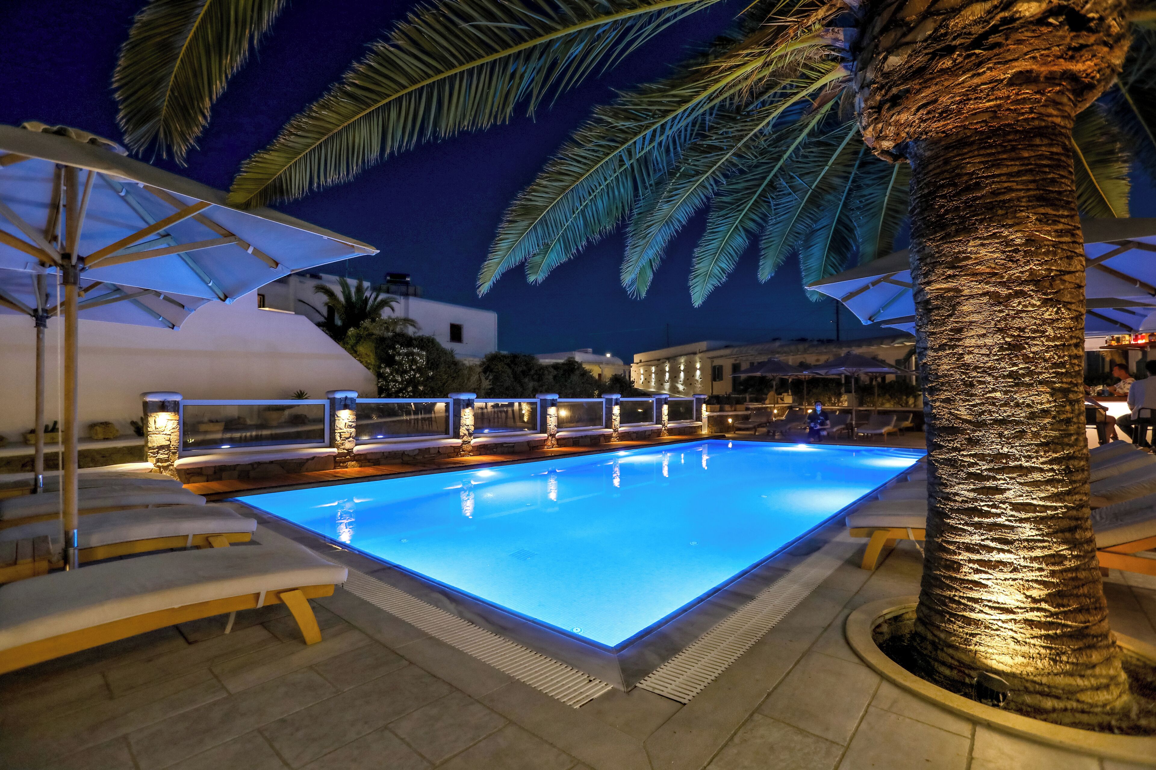 A swimming pool with lounge chairs and umbrellas, surrounded by palm trees and a night sky.