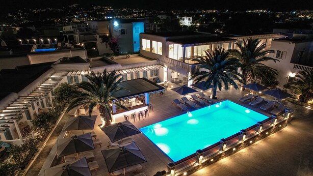 Uma área de piscina no terraço com espreguiçadeiras e guarda-sóis, cercada por palmeiras e um edifício com grandes janelas.