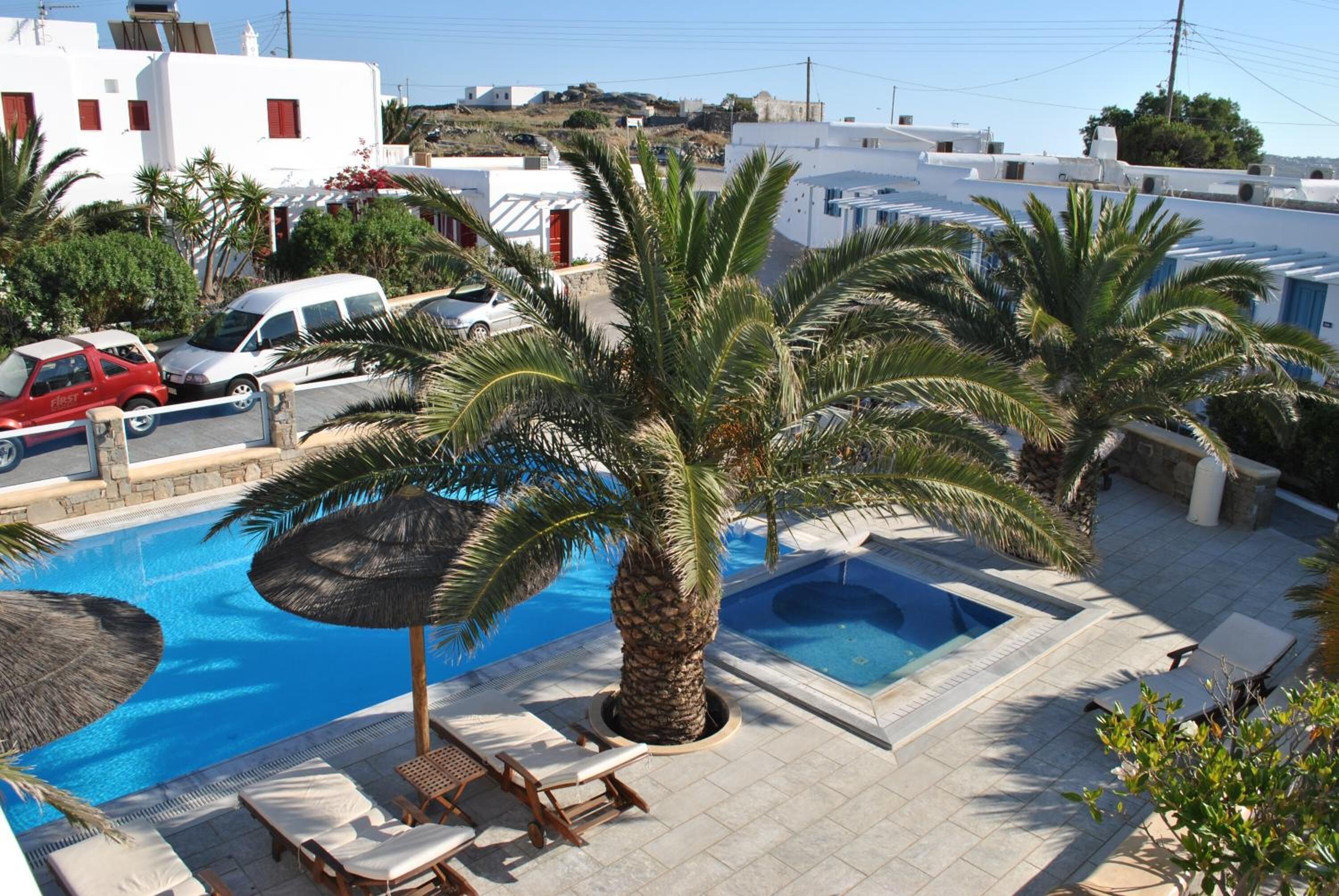 Zona de piscina con sillones de descanso, palmeras y vistas a edificios blancos al fondo.