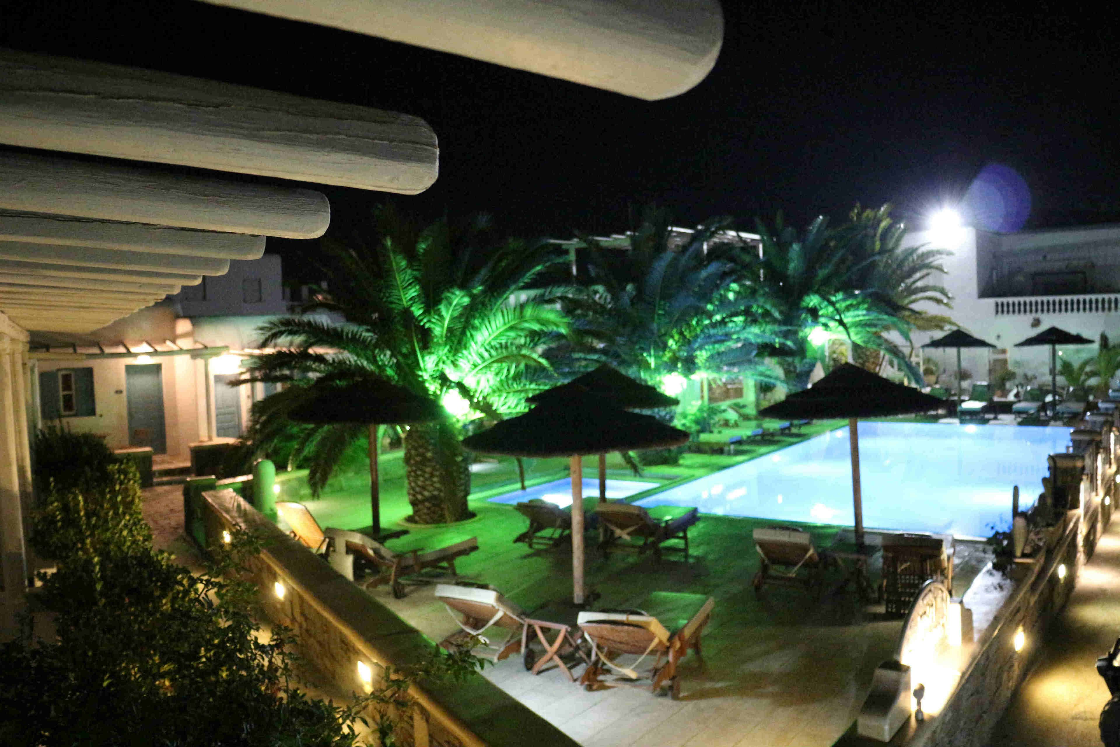 A nighttime pool area with lounge chairs and umbrellas, surrounded by palm trees and a building in the background.