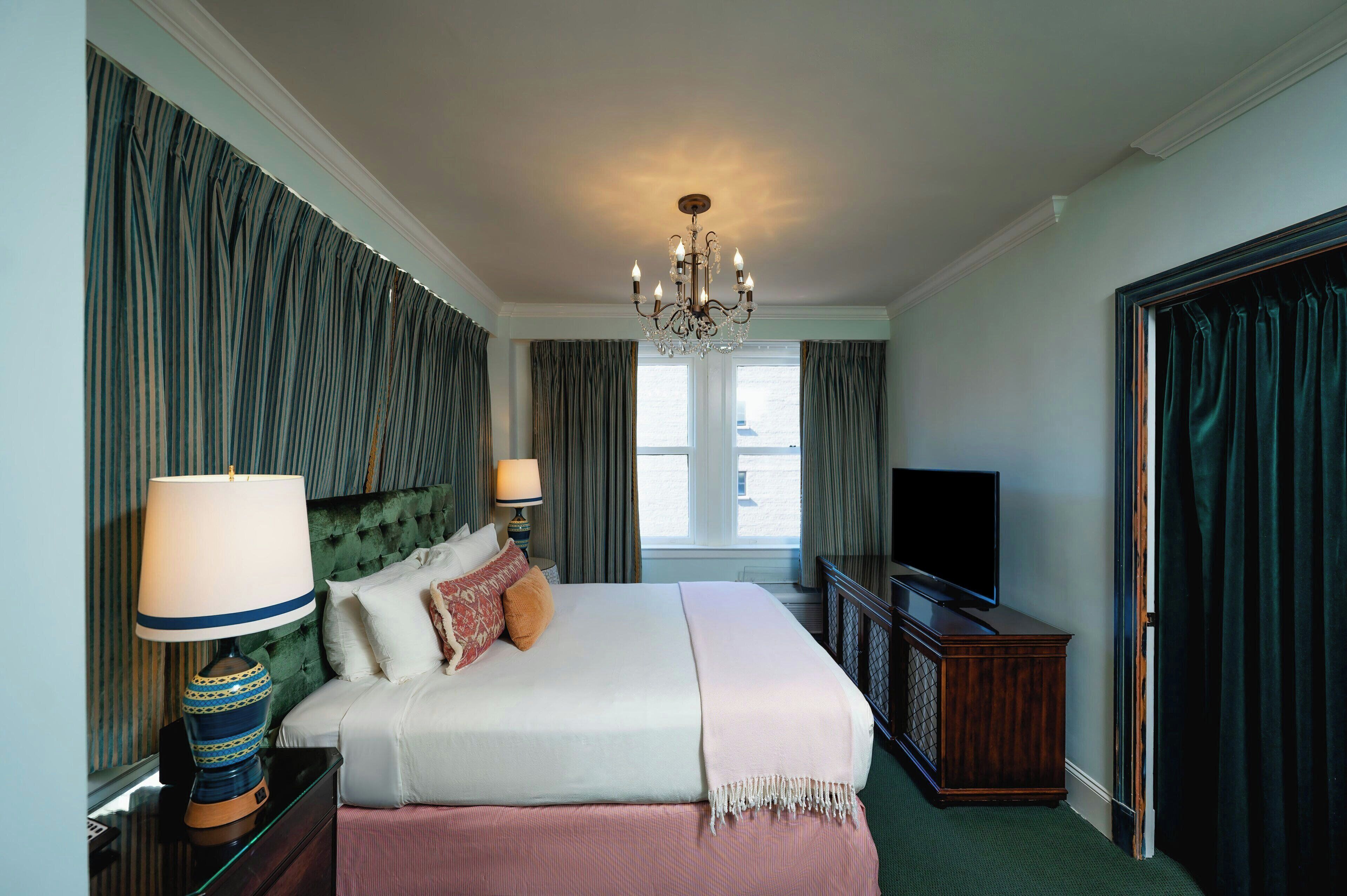 Une chambre d’hôtel avec un lit, deux lampes de chevet, une télévision, une commode et un lustre.