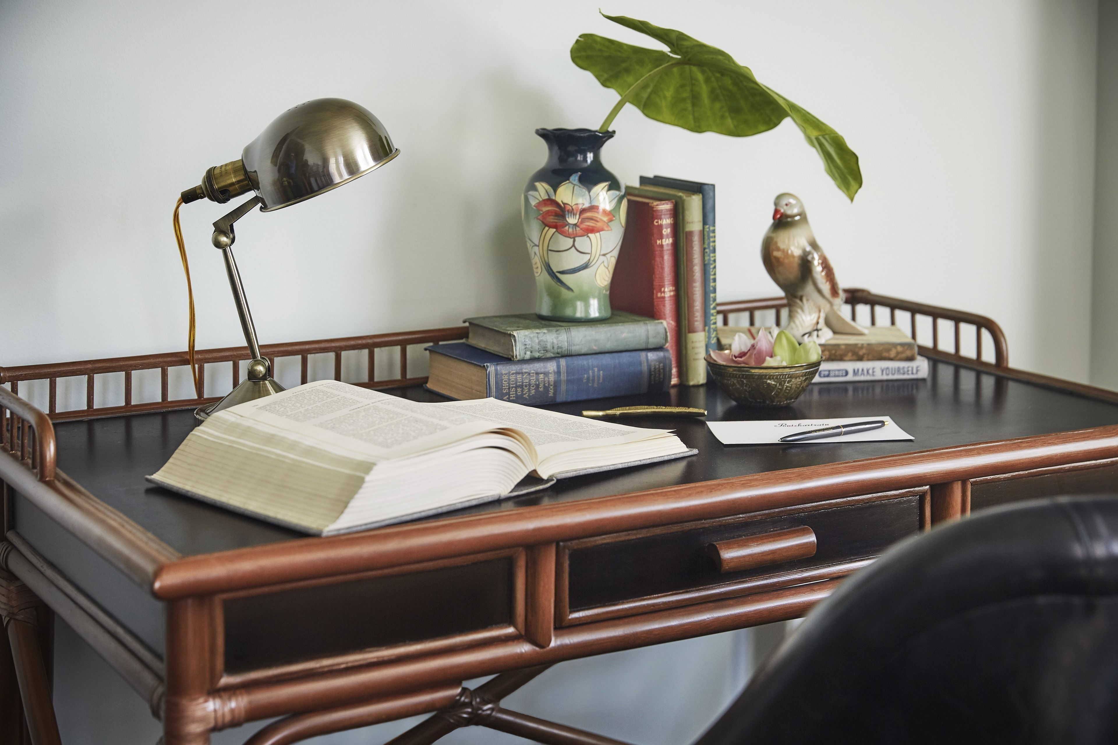 A wooden desk with an open book, a lamp, a vase with a plant, and a bird statue.