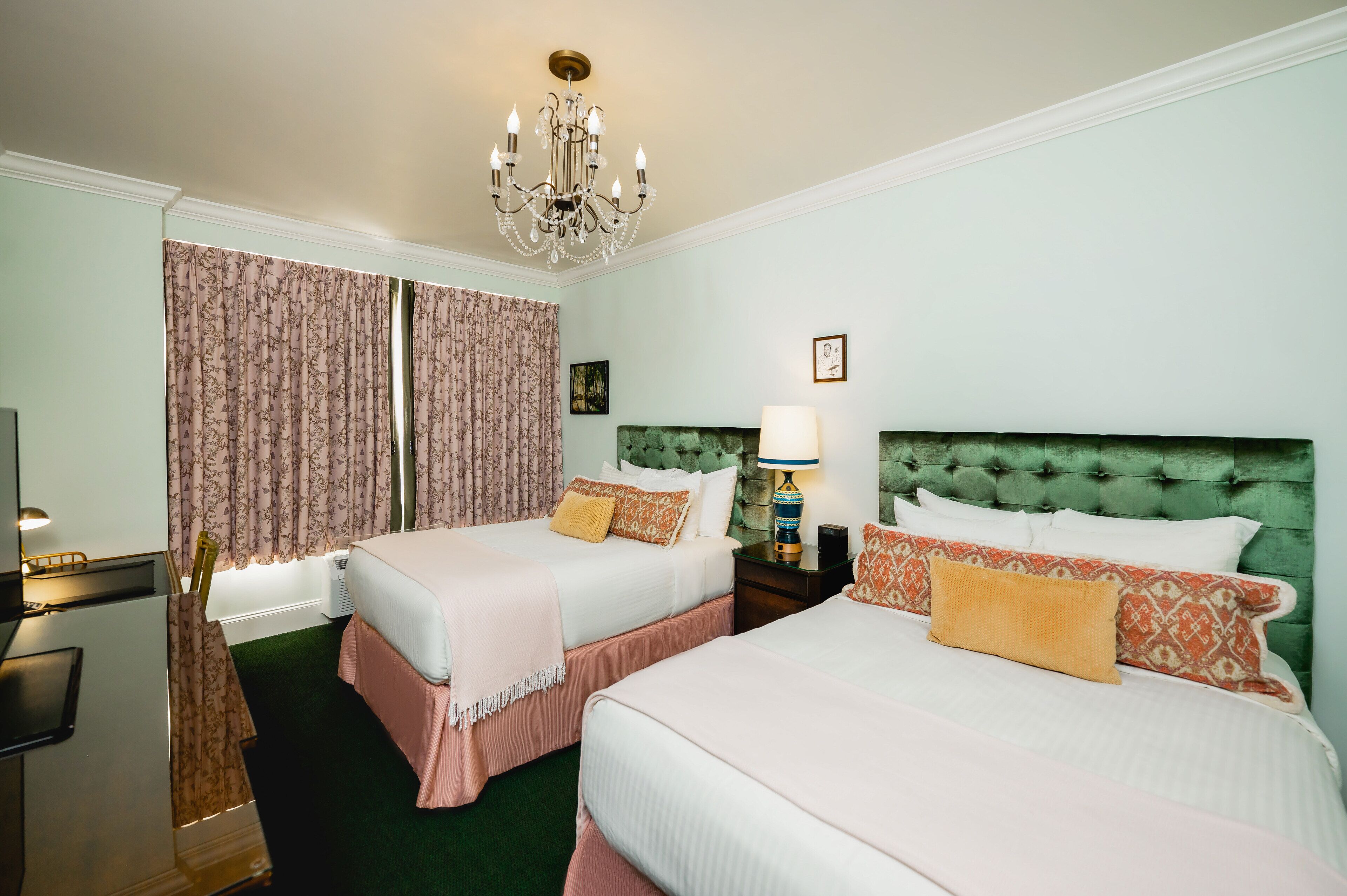A hotel room with two beds, a chandelier, a green upholstered headboard, and a nightstand with a lamp.