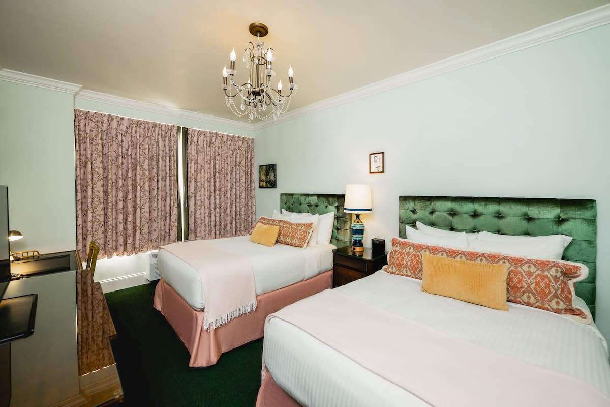 A hotel room with two beds, a chandelier, a green upholstered headboard, and a nightstand with a lamp.