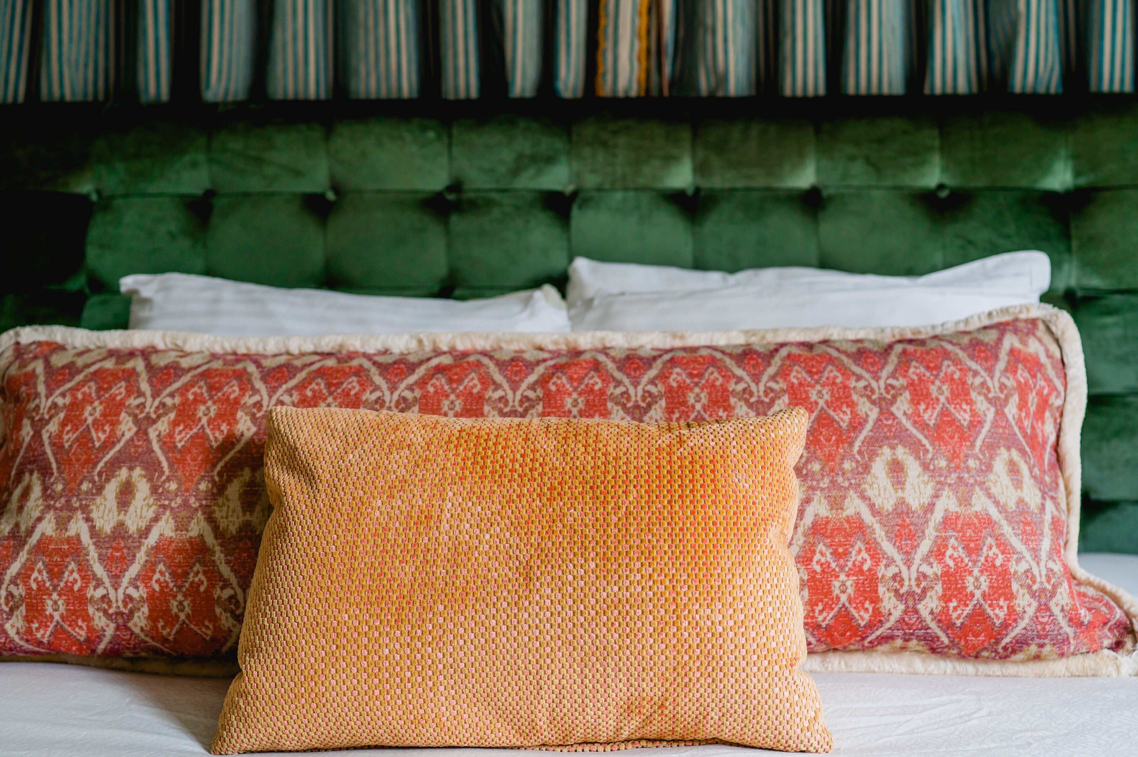 Una cama con cabecera tapizada en verde, dos almohadas estampadas y una almohada naranja con textura.