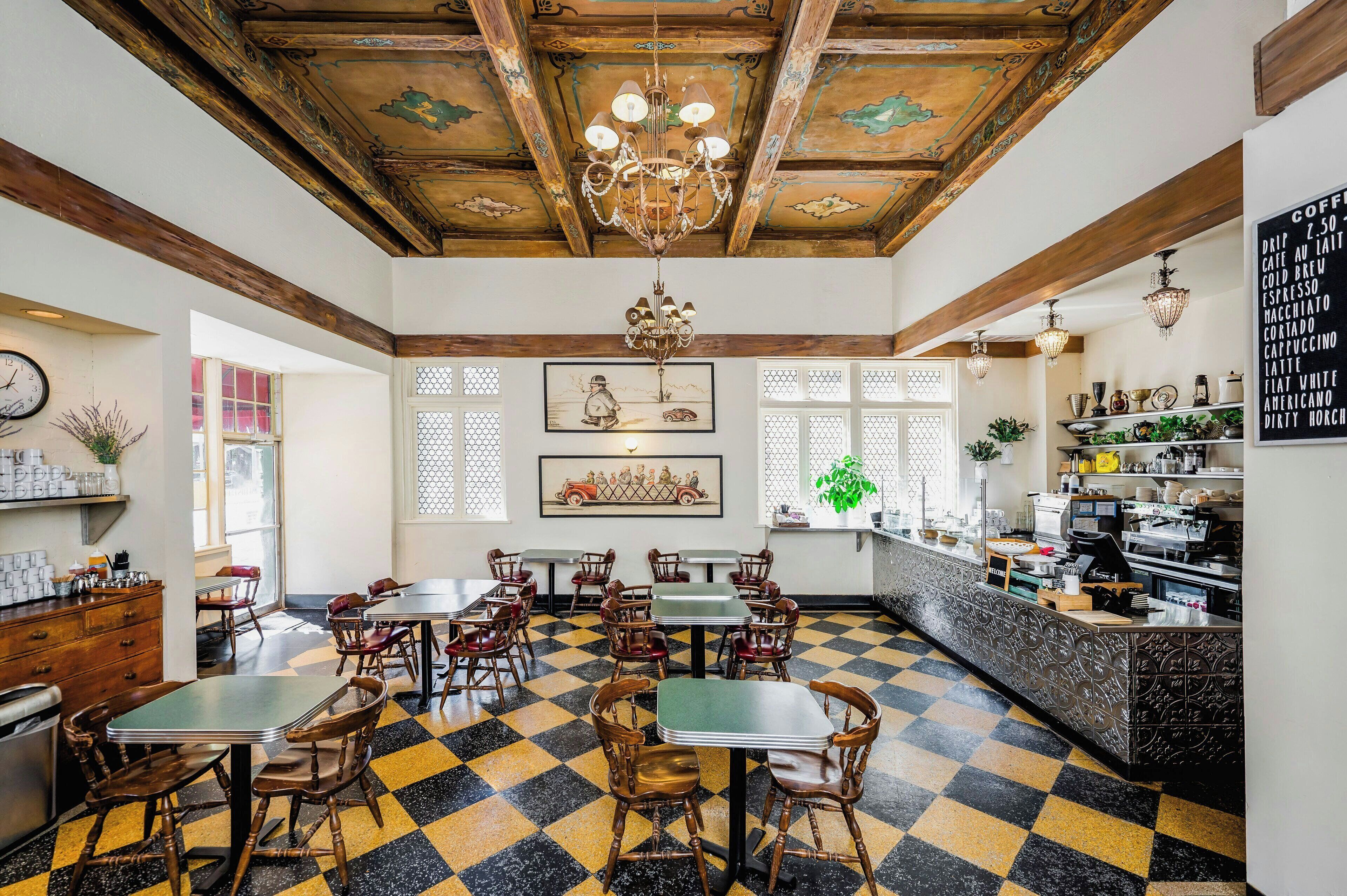 A cafe with checkered flooring, wooden chairs, tables, a bar area, and a menu board.