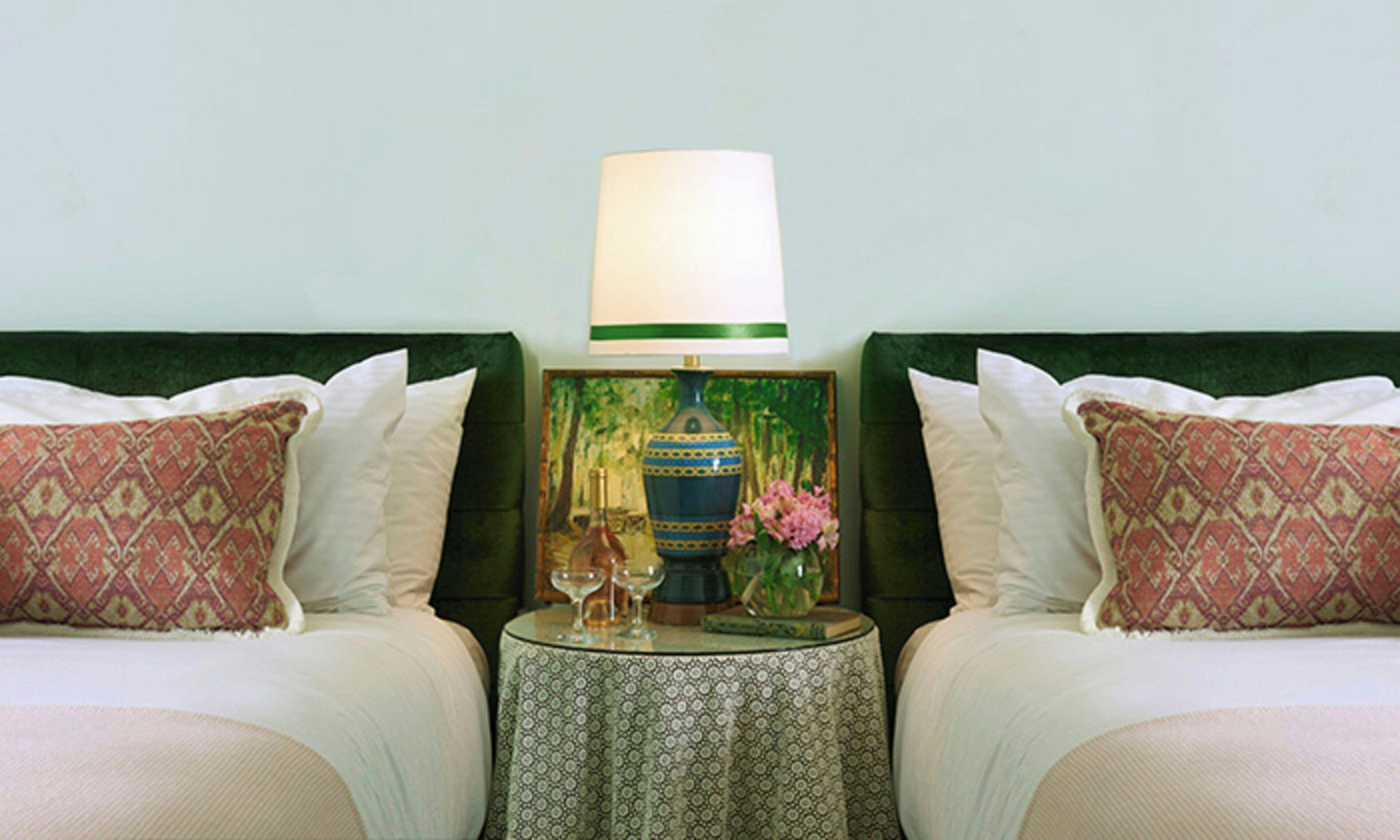 Habitación de hotel con dos camas, cada una con almohadas estampadas, una mesita redonda con una lámpara y un cuadro en la pared.