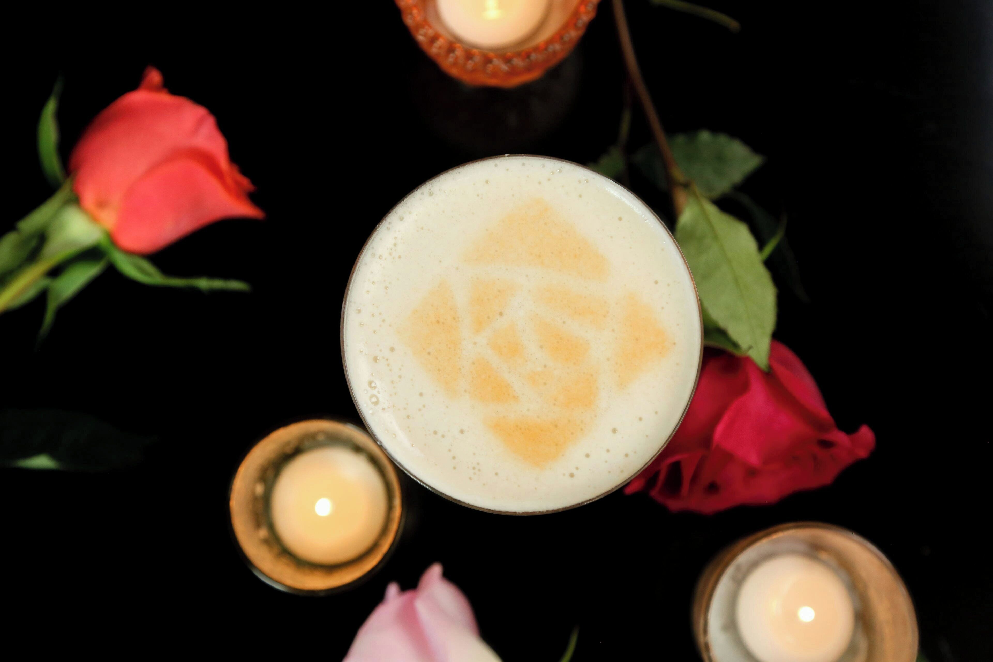 A glass with a frothy beverage, surrounded by candles and flowers on a reflective surface.