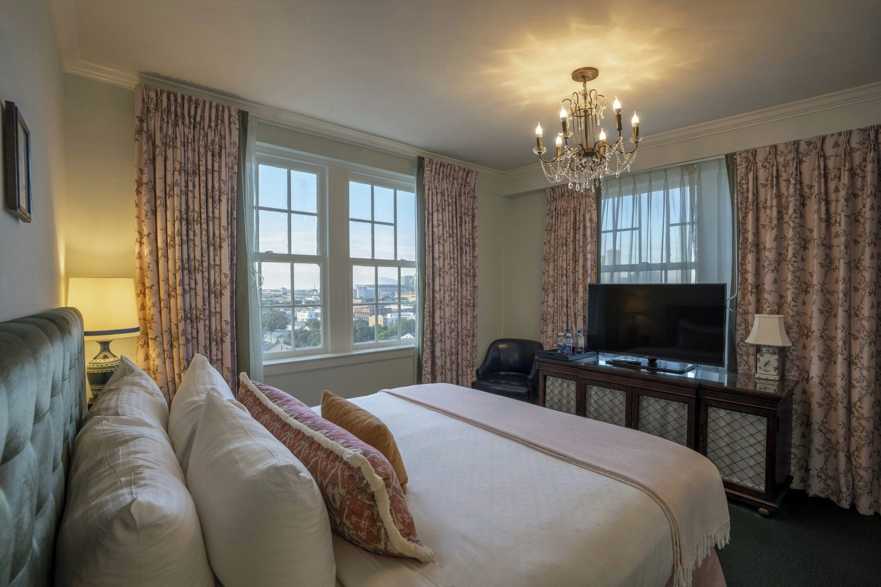 A hotel room with a large bed, a television, a desk with a lamp, and a view of the city through the windows.