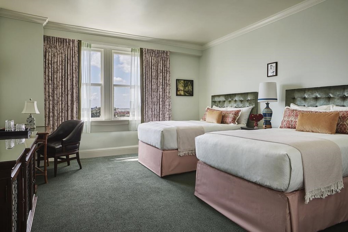 A hotel room with two beds, a desk with a chair, and a window with curtains.