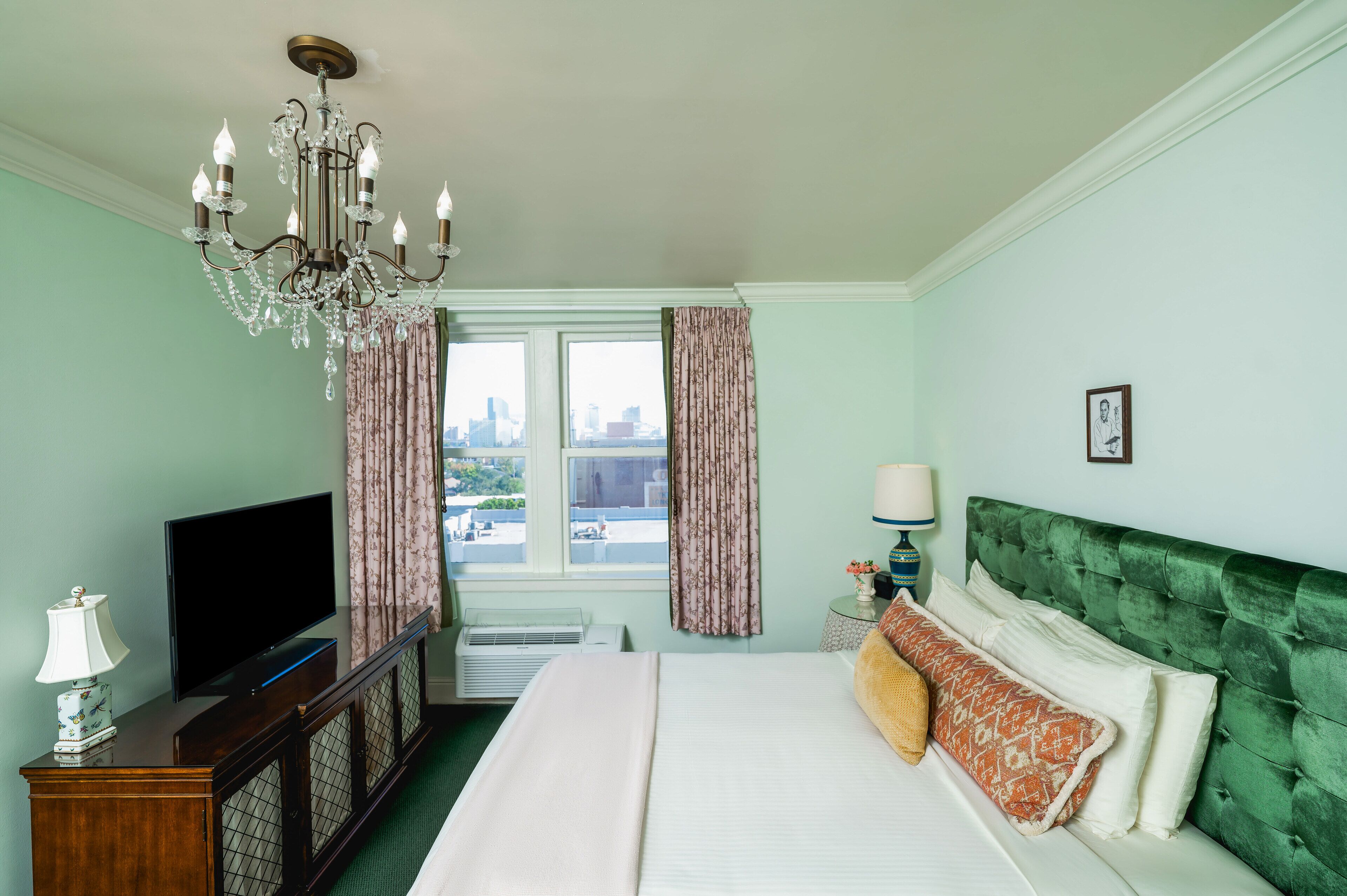 A hotel room with a bed, a television, a chandelier, a window with curtains, and a city view.