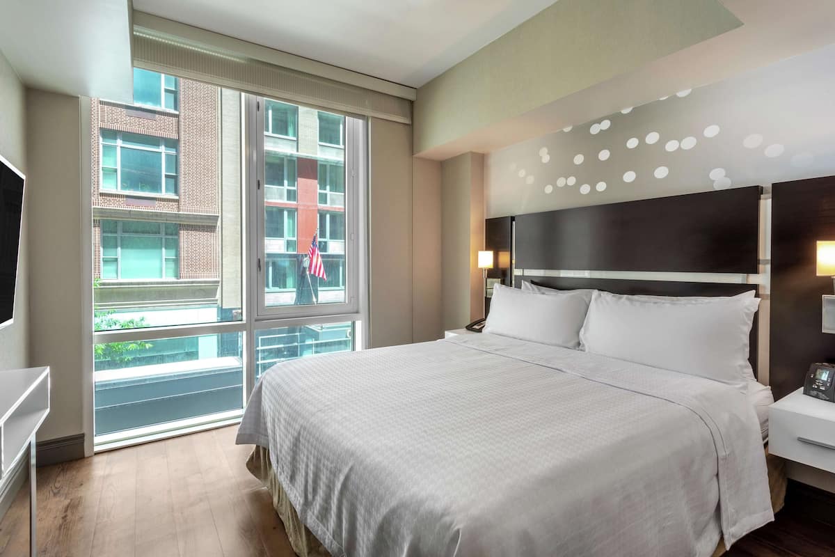 Ein Hotelzimmer mit einem großen Bett, einem Fernseher, einem Nachttisch und einem Fenster mit Blick auf ein Gebäude und eine Flagge.