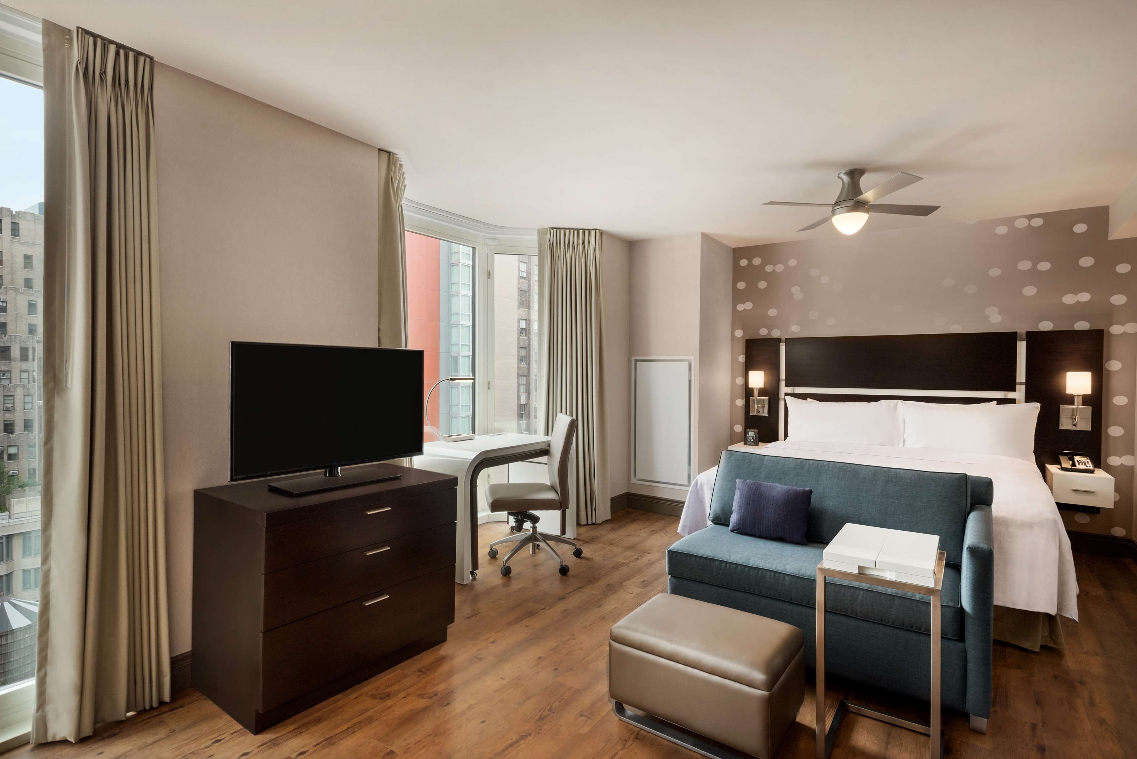 A hotel room with a bed, a desk, a TV, a chair, a sofa, and a ceiling fan.