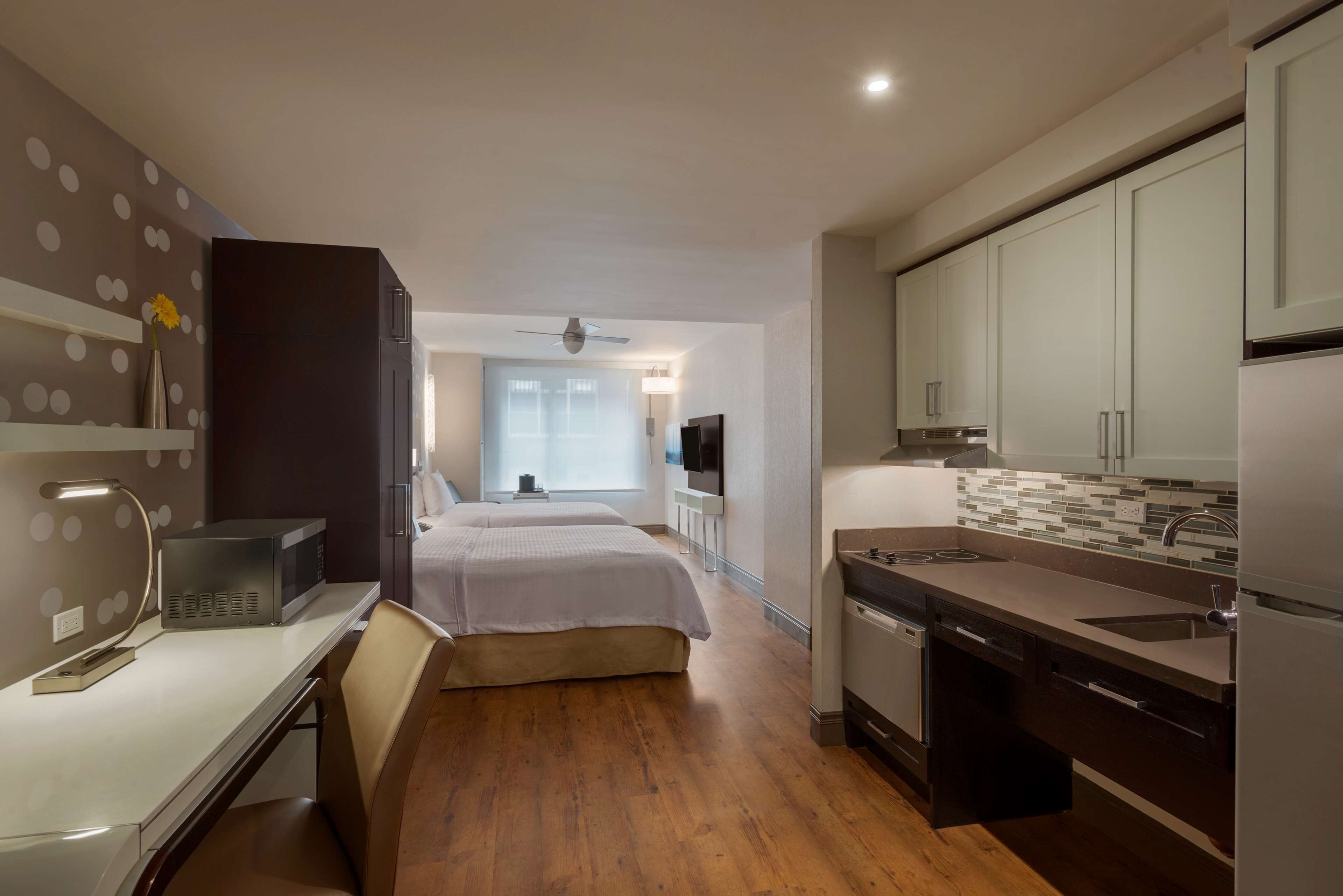 Une chambre d’hôtel moderne avec un plancher en bois, un lit, un bureau avec une lampe, une télévision, un coin cuisine avec des placards et un évier, et une vue sur le couloir.