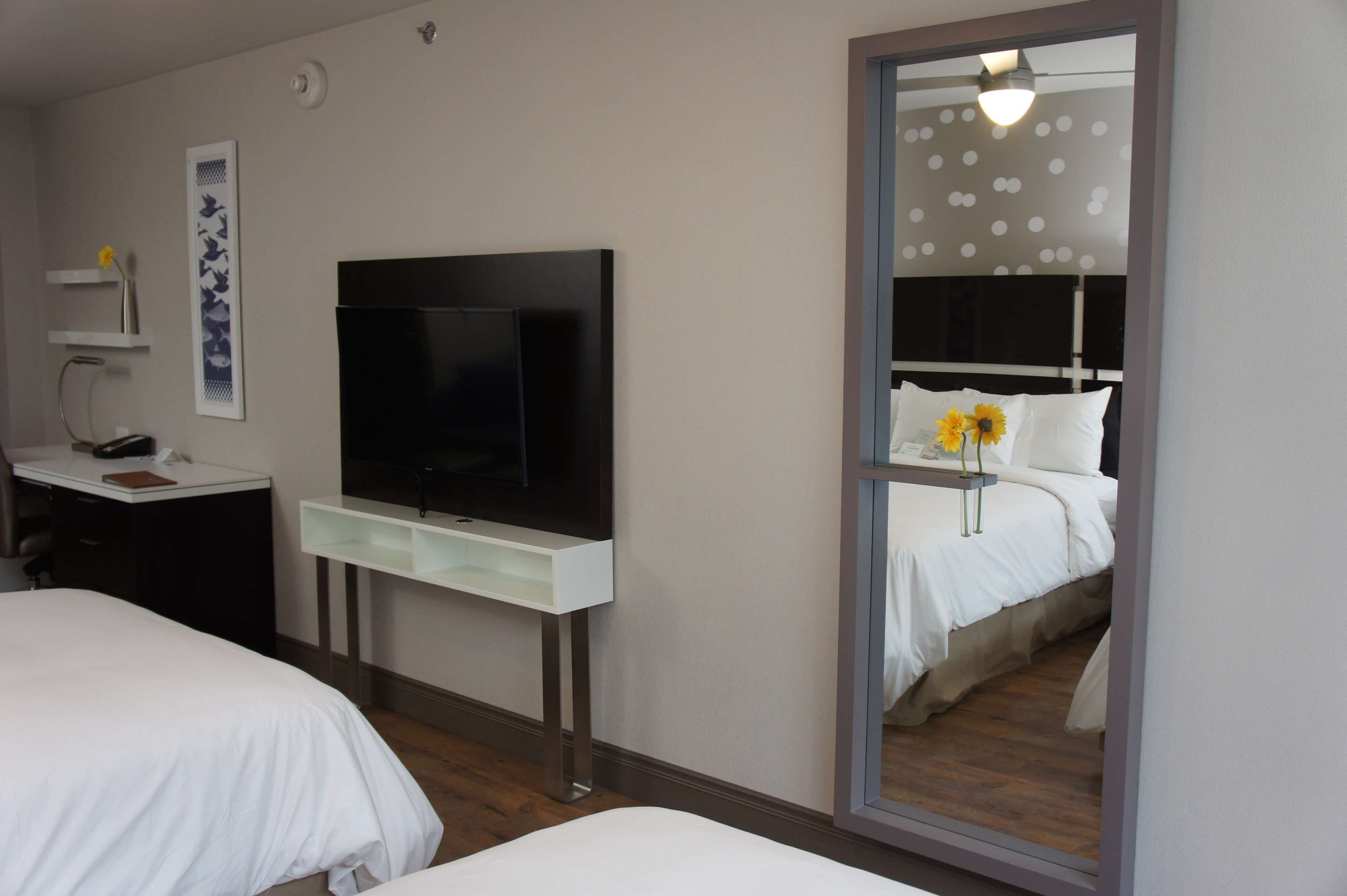 A hotel room with a bed, a desk with a flat-screen TV, a mirror, and a wall-mounted shelf with a vase.