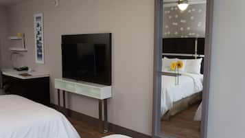 A hotel room with a bed, a desk with a flat-screen TV, a mirror, and a wall-mounted shelf with a vase.