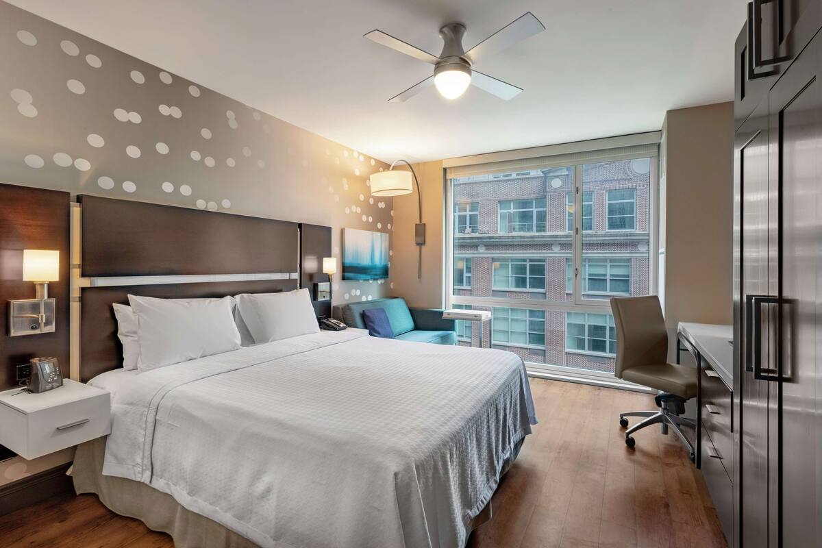 Ein Hotelzimmer mit einem großen Bett, einem Schreibtisch, einem Stuhl, einem Deckenventilator und einem Fenster mit Blick auf Gebäude.