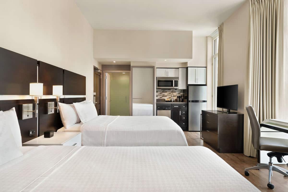 Ein Hotelzimmer mit einem großen Bett, Nachttischen, einem Schreibtisch mit Stuhl, einem Fernseher und einer Küchenzeile mit Mikrowelle und Kühlschrank.