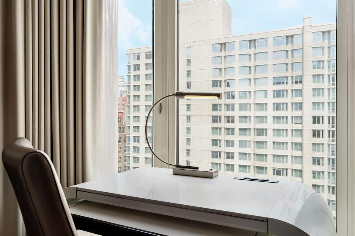 Ein moderner Schreibtisch mit Lampe, einem Stuhl und Blick auf hohe Gebäude durch ein Fenster.