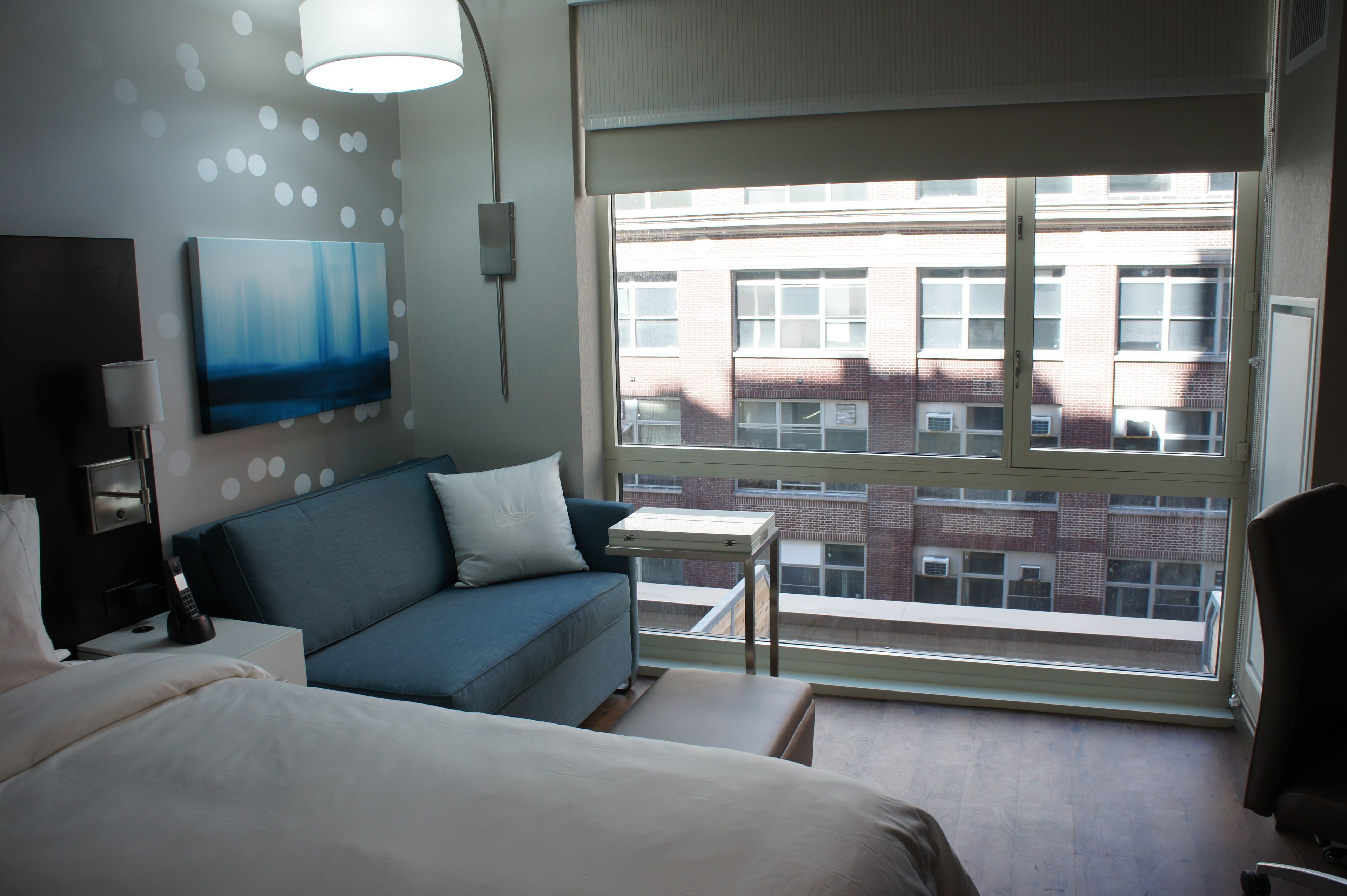 A modern hotel room with a large window, a blue sofa, a bedside table, and a painting on the wall.