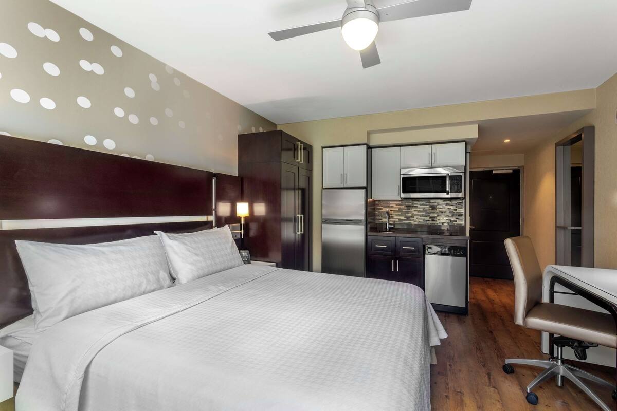 Ein Hotelzimmer mit Bett, Schreibtisch, Stuhl, Küchenzeile und Deckenventilator.