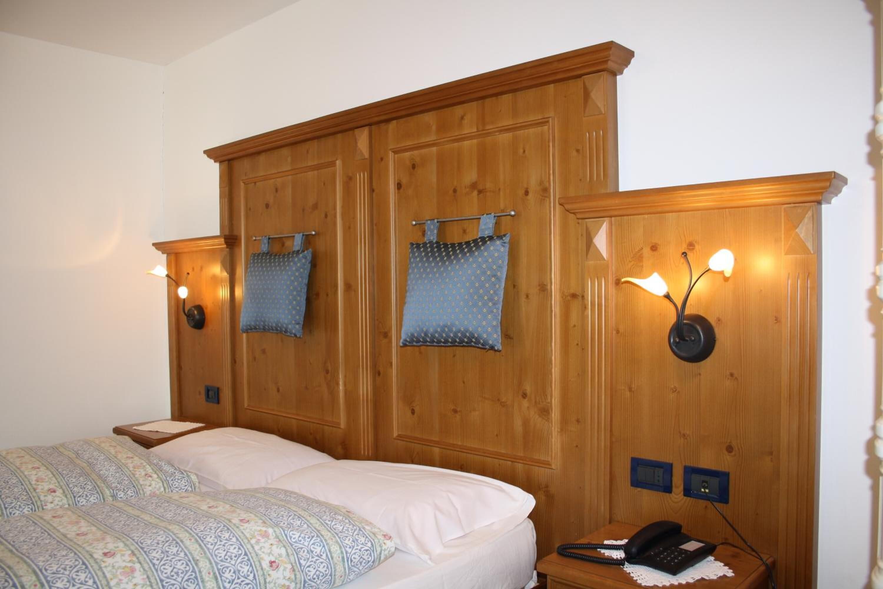 A bedroom with a wooden headboard, a bed with patterned bedding, two wall-mounted lamps, and a bedside telephone.