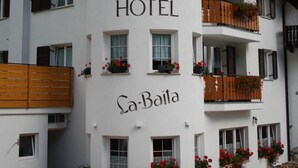 Un hotel chiamato "La-Baita" con facciata bianca, numerosi balconi fioriti di rosso e uno sfondo montuoso.