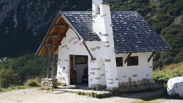 Un piccolo edificio in pietra con tetto spiovente, una porta di legno e un cartello con su scritto 
