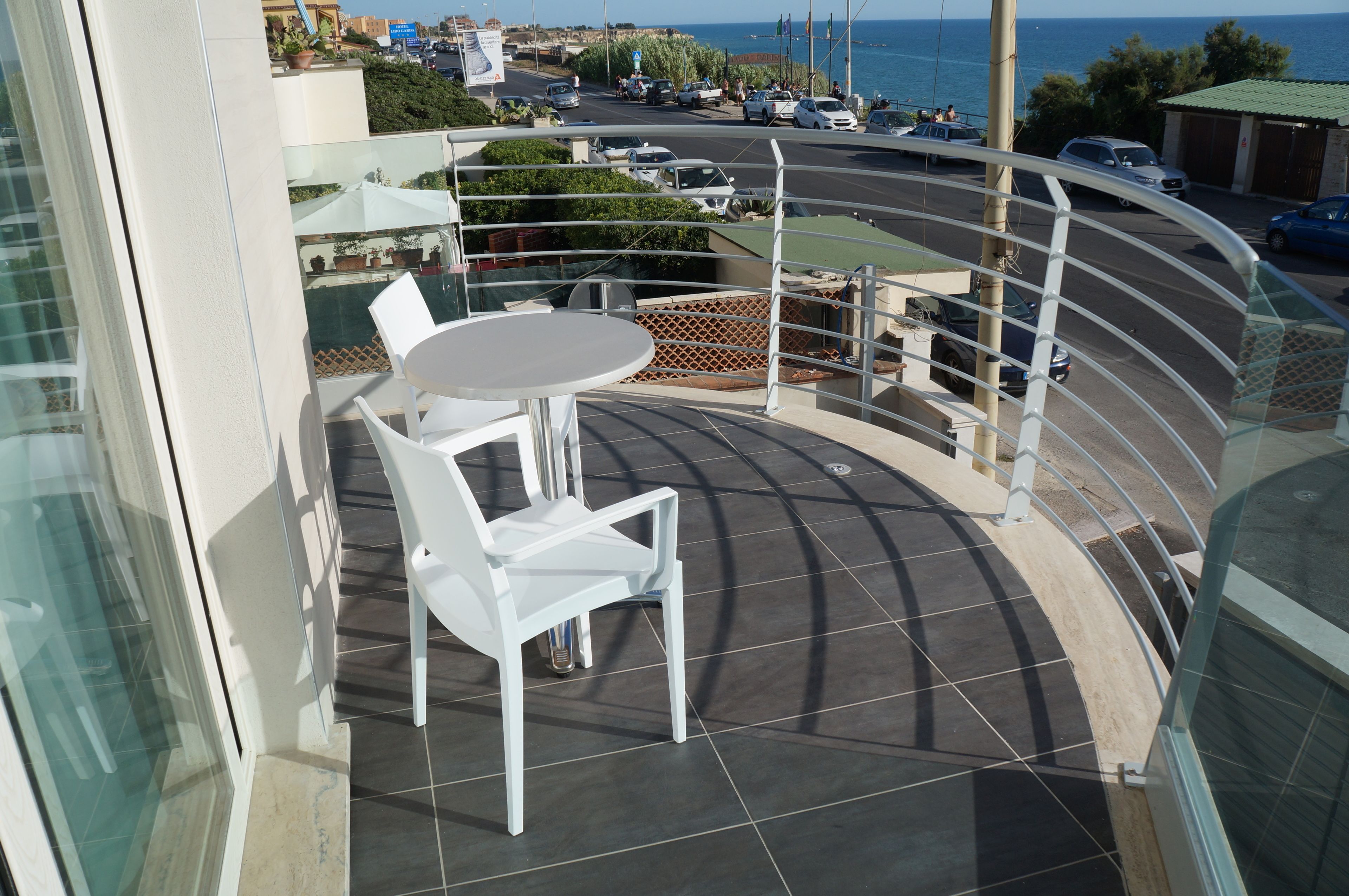 Ein Balkon mit Tisch und Stühlen, der auf eine Straße mit geparkten Autos und Meerblick blickt.