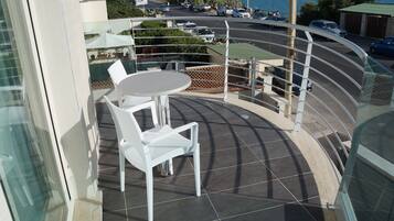 Ein Balkon mit Tisch und Stühlen, der auf eine Straße mit geparkten Autos und Meerblick blickt.