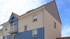 Edifici residencial de dues plantes amb façana blava i beix, un balcó al segon pis i un carrer d'accés reixat.