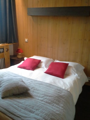 A bed with white bedding and two red pillows, a wooden headboard, and a small table with a red lamp. - Hôtel Val Joly (Saint-Gervais-les-Bains)