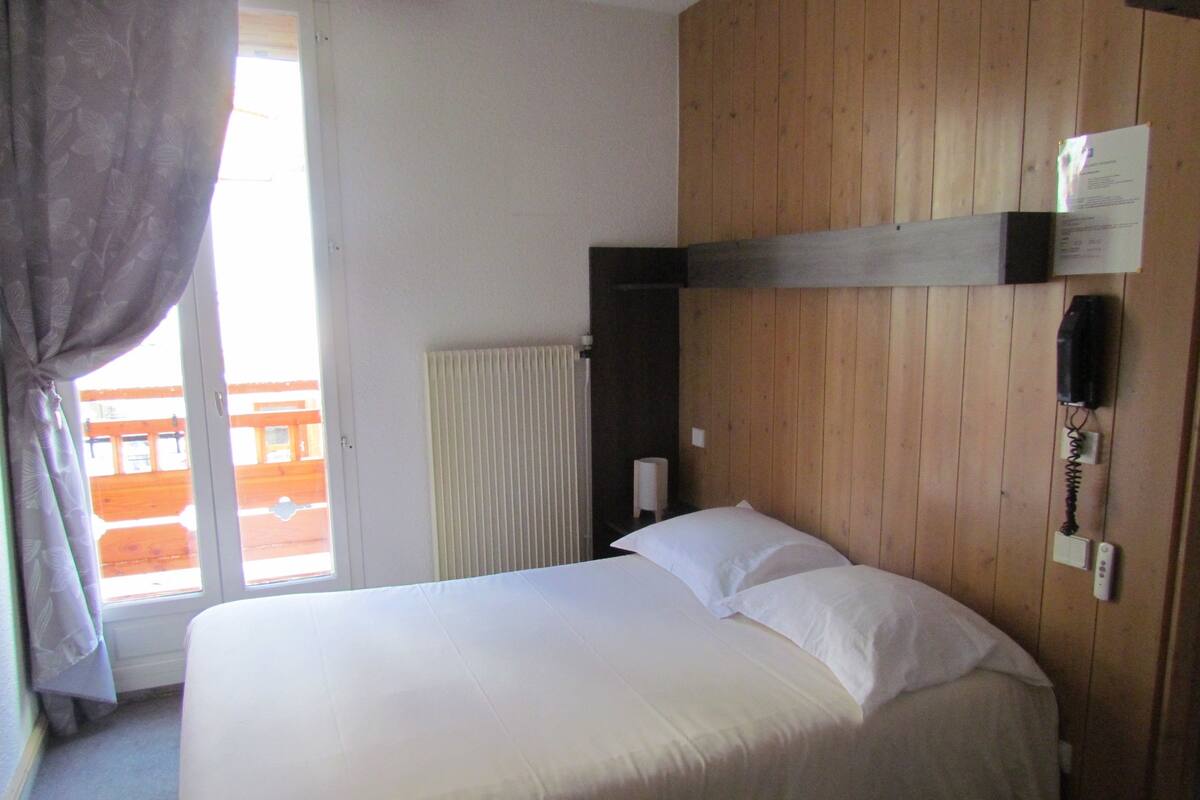 Une chambre d’hôtel avec un lit, une fenêtre avec des rideaux, un radiateur et un mur en bois.
