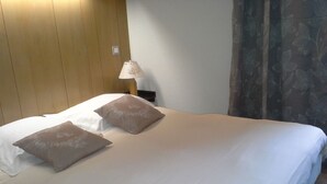 A neatly made bed with two pillows, a wooden headboard, a bedside lamp, and a curtain with a floral pattern. - Hôtel Val Joly (Saint-Gervais-les-Bains)
