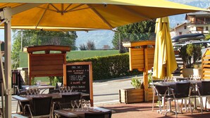 An outdoor dining area with tables and chairs under a yellow umbrella, a menu board, and a mountainous backdrop. - Hôtel Val Joly (Saint-Gervais-les-Bains)