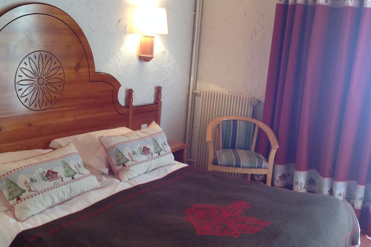 Un dormitorio con cabecera de madera, cama con dos almohadas, lámpara de noche, una silla y una ventana con cortinas.