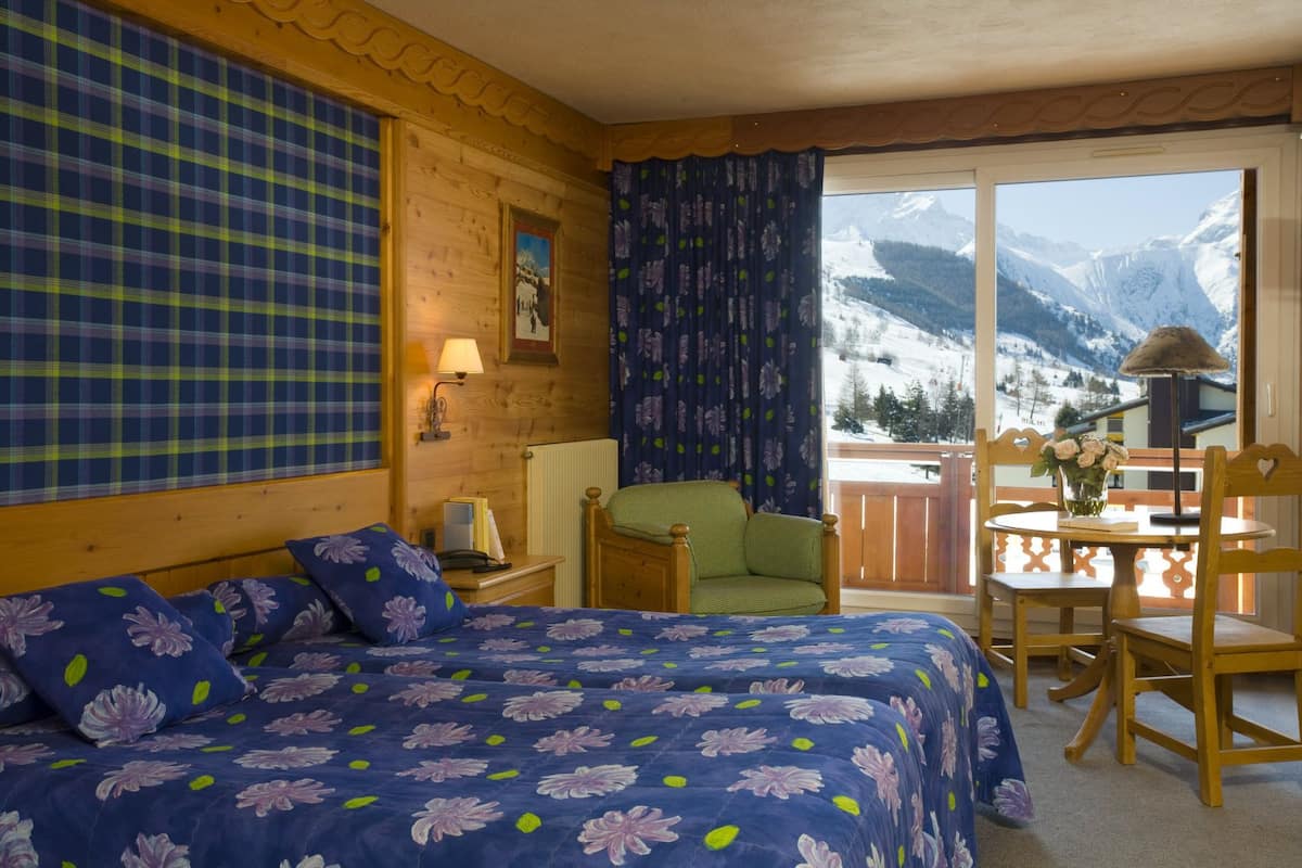Un dormitorio con cama, una silla, una mesa con flores y vistas a montañas nevadas.