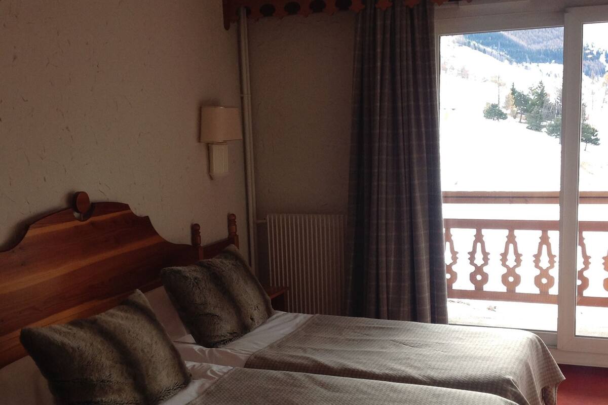 Habitación de hotel con dos camas, cabecero de madera, ventana con vistas a montañas y balcón con barandilla.