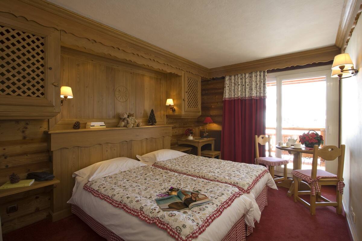 Habitación acogedora con dos camas, una mesita y una silla. Objetos decorativos en las estanterías y una ventana con cortinas.