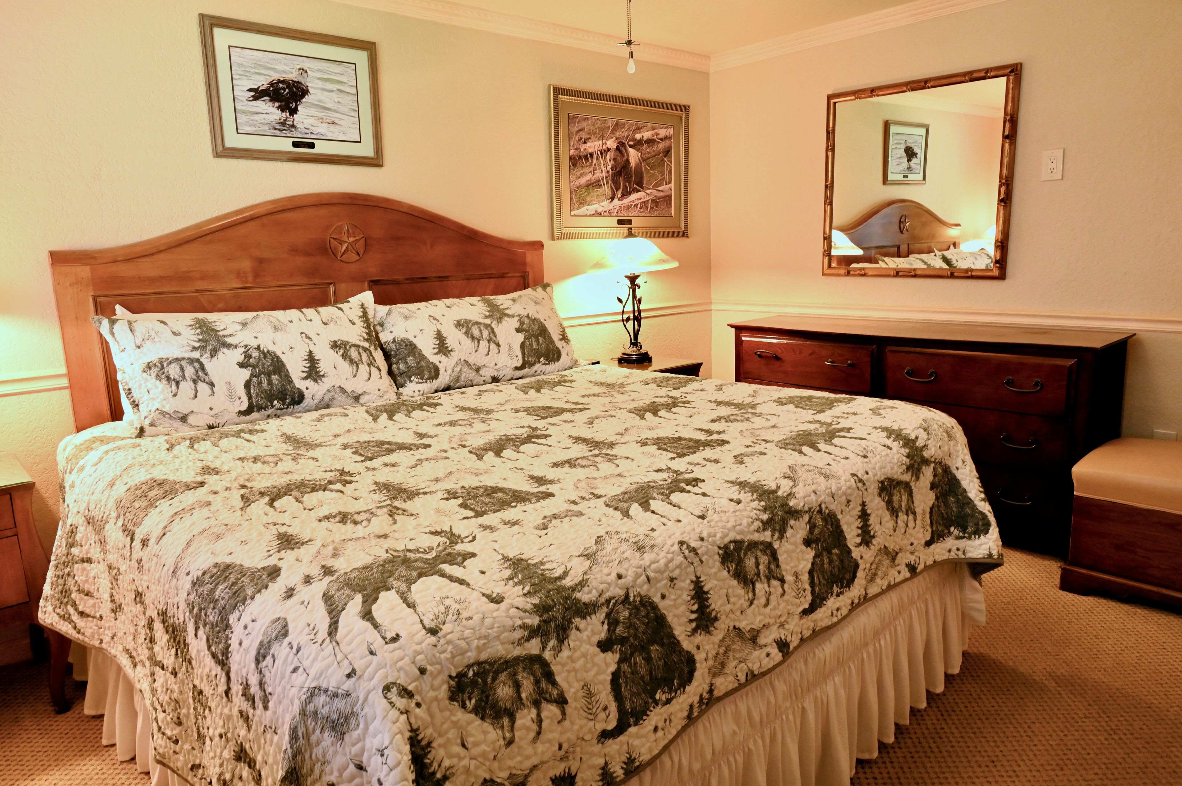 A bedroom with a large bed, a nightstand, a dresser, a lamp, and two framed pictures on the wall.
