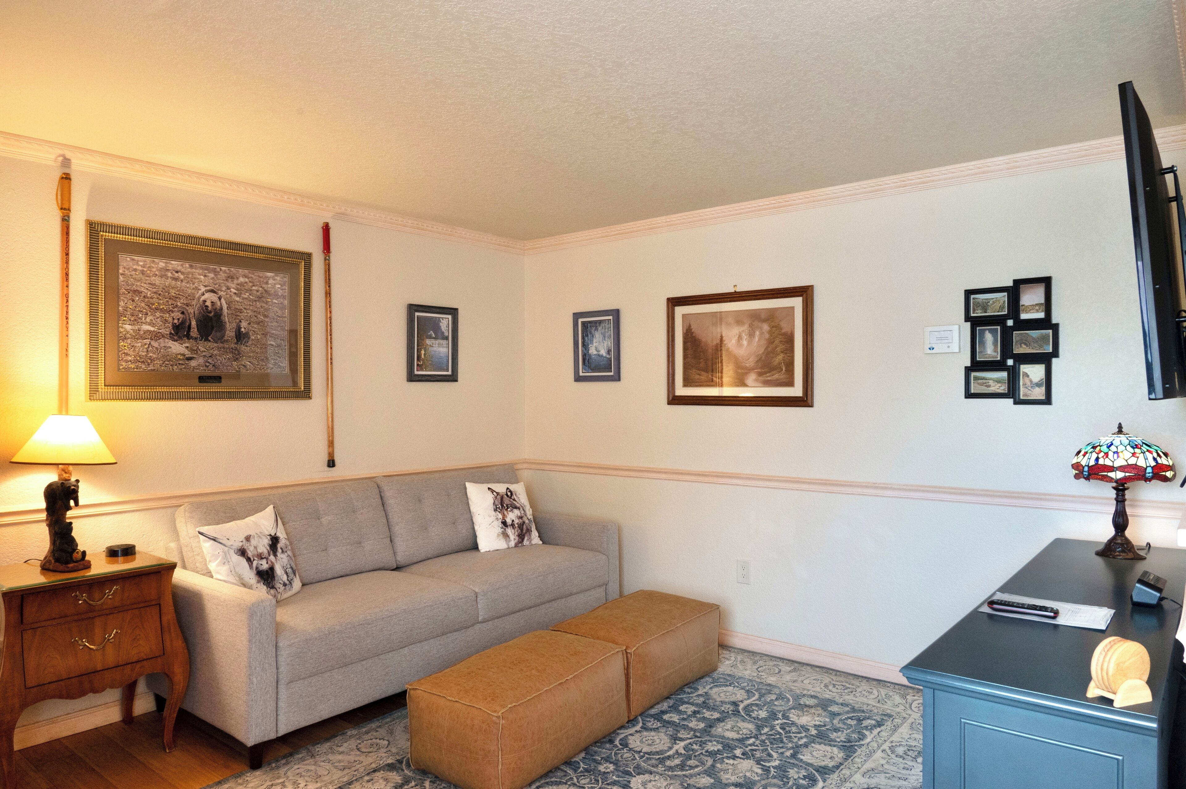 A living room with a sofa, ottoman, and a TV on a stand. There are framed pictures on the wall and a lamp on a side table.