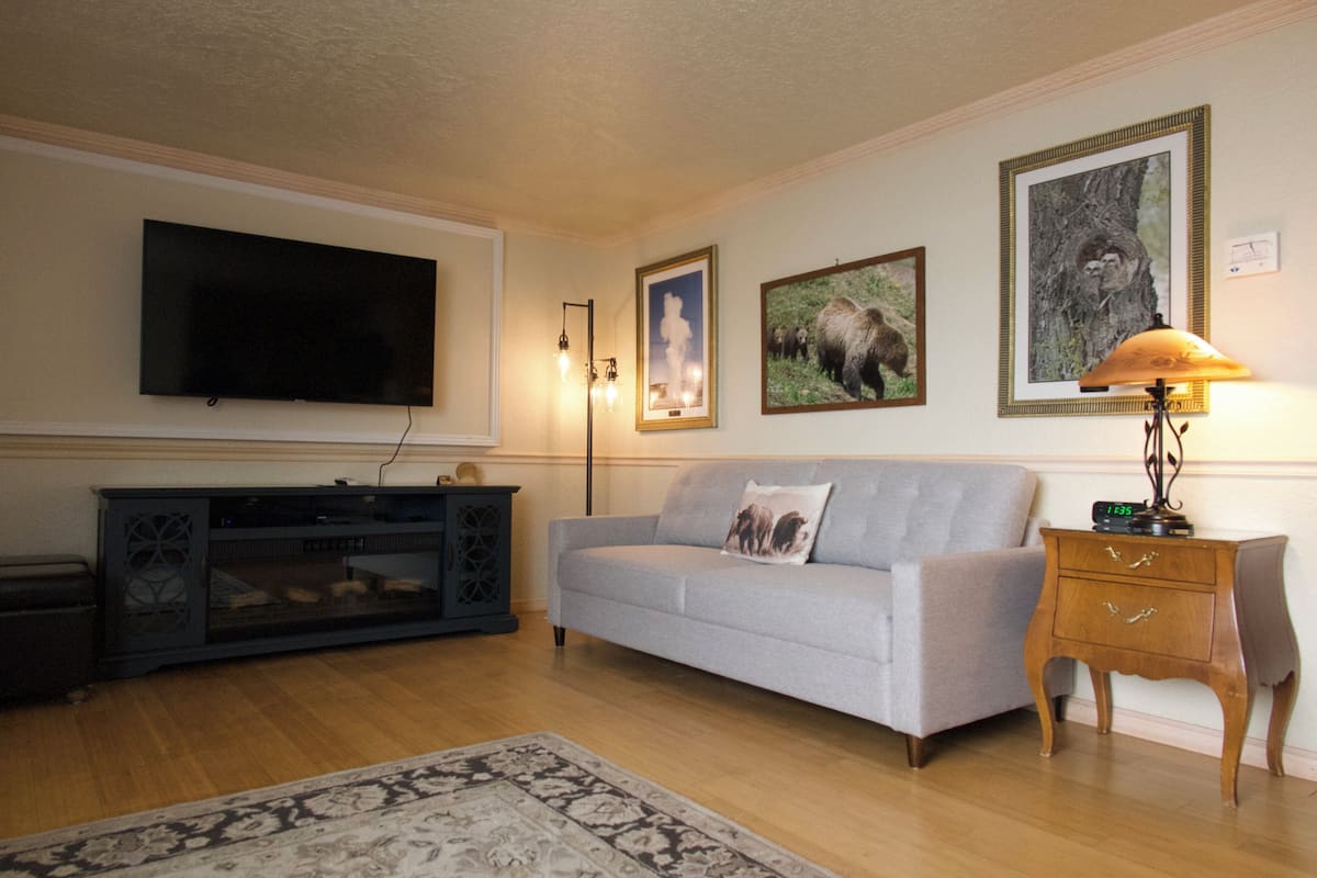A living room with a flat-screen TV mounted on the wall, a grey sofa with a decorative pillow, a wooden side table with a lamp, and framed pictures on the wall.