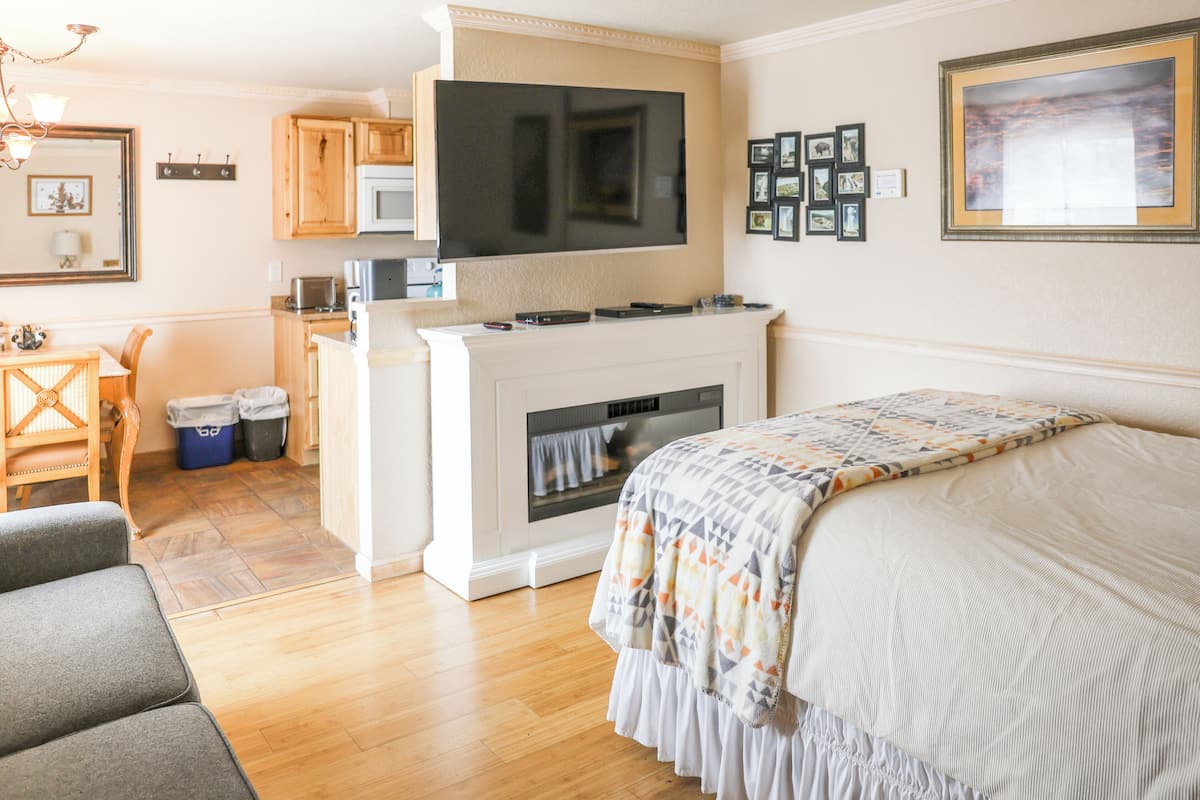A modern, well-lit room with a large bed, a flat-screen TV mounted above a fireplace, wooden cabinets, and a dining area with a table and chairs.